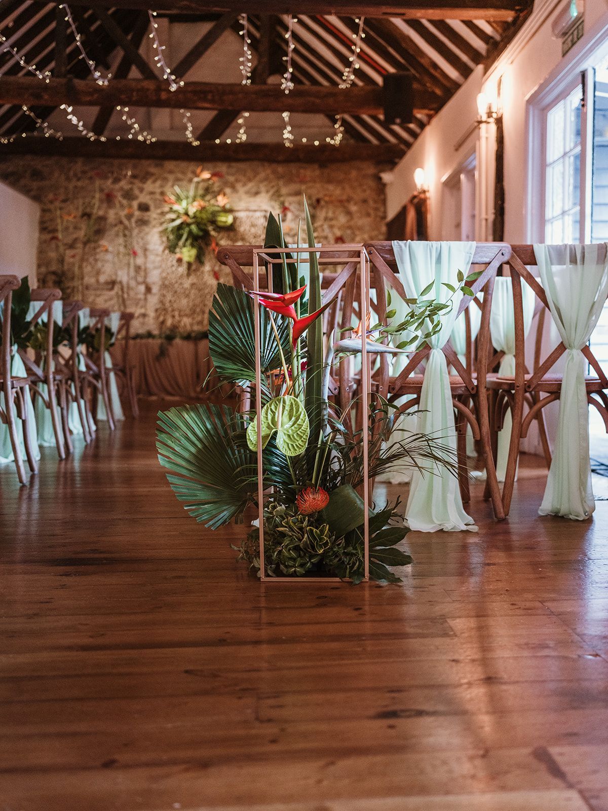 Rustic indoor wedding aisle with floral arrangements, wooden flooring, and tall windows letting in soft light