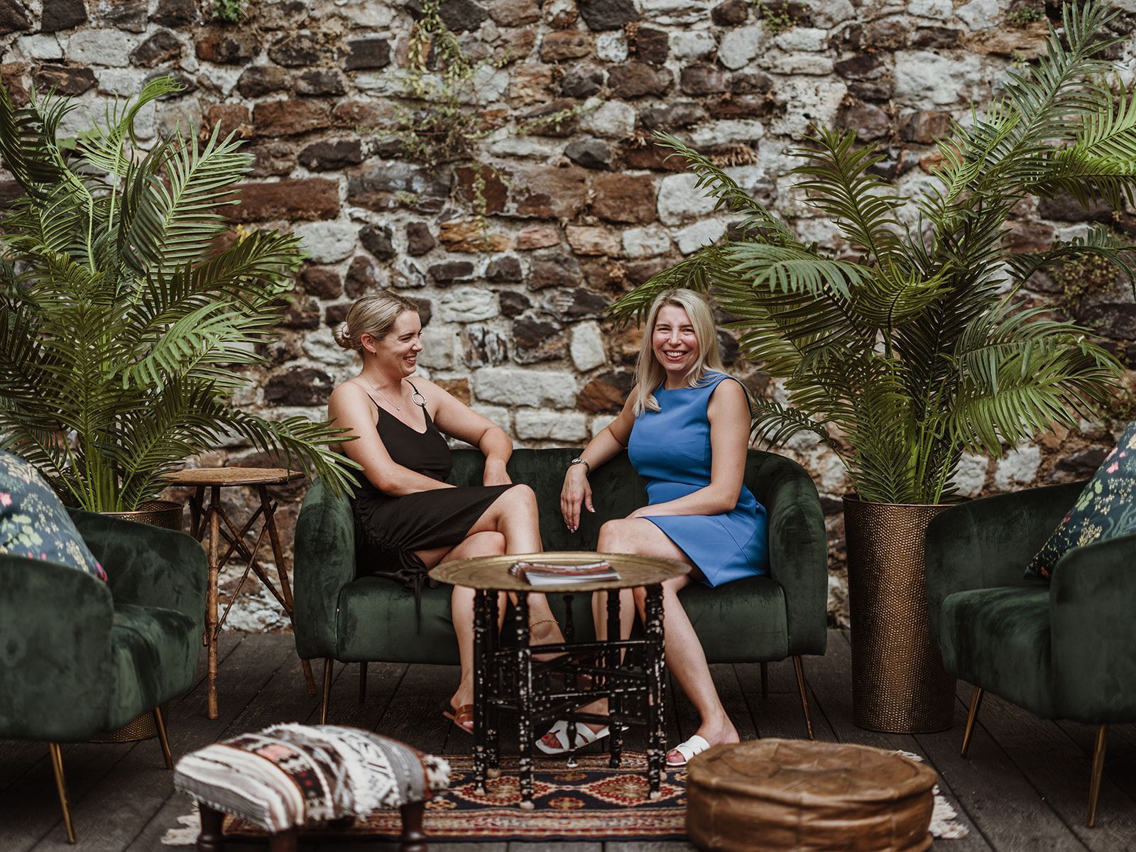Two women sit on outdoor chairs beside a small table against a stone wall and potted palms.