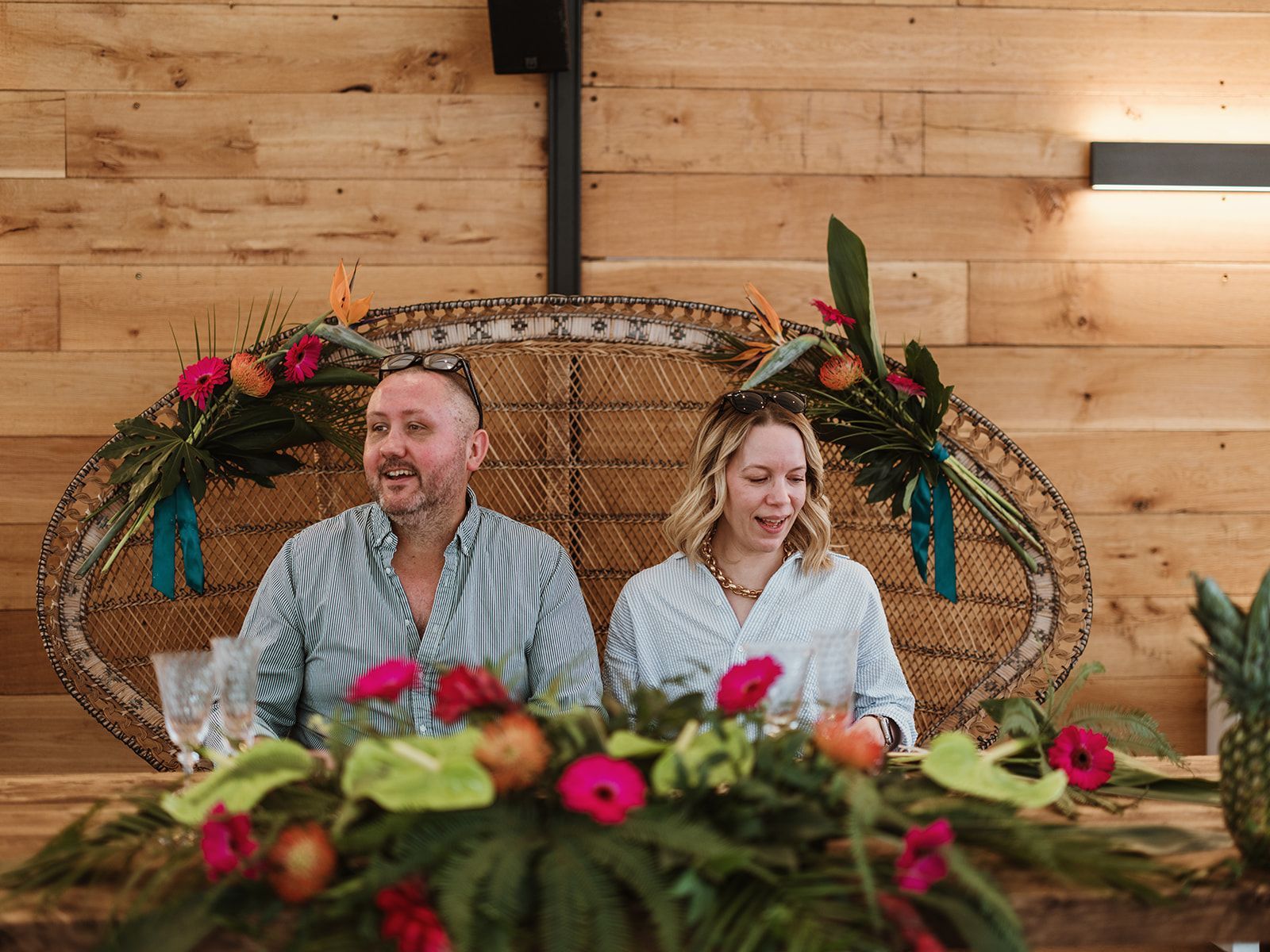 Two people seated at a decorated wooden table with floral arrangements in a rustic indoor setting