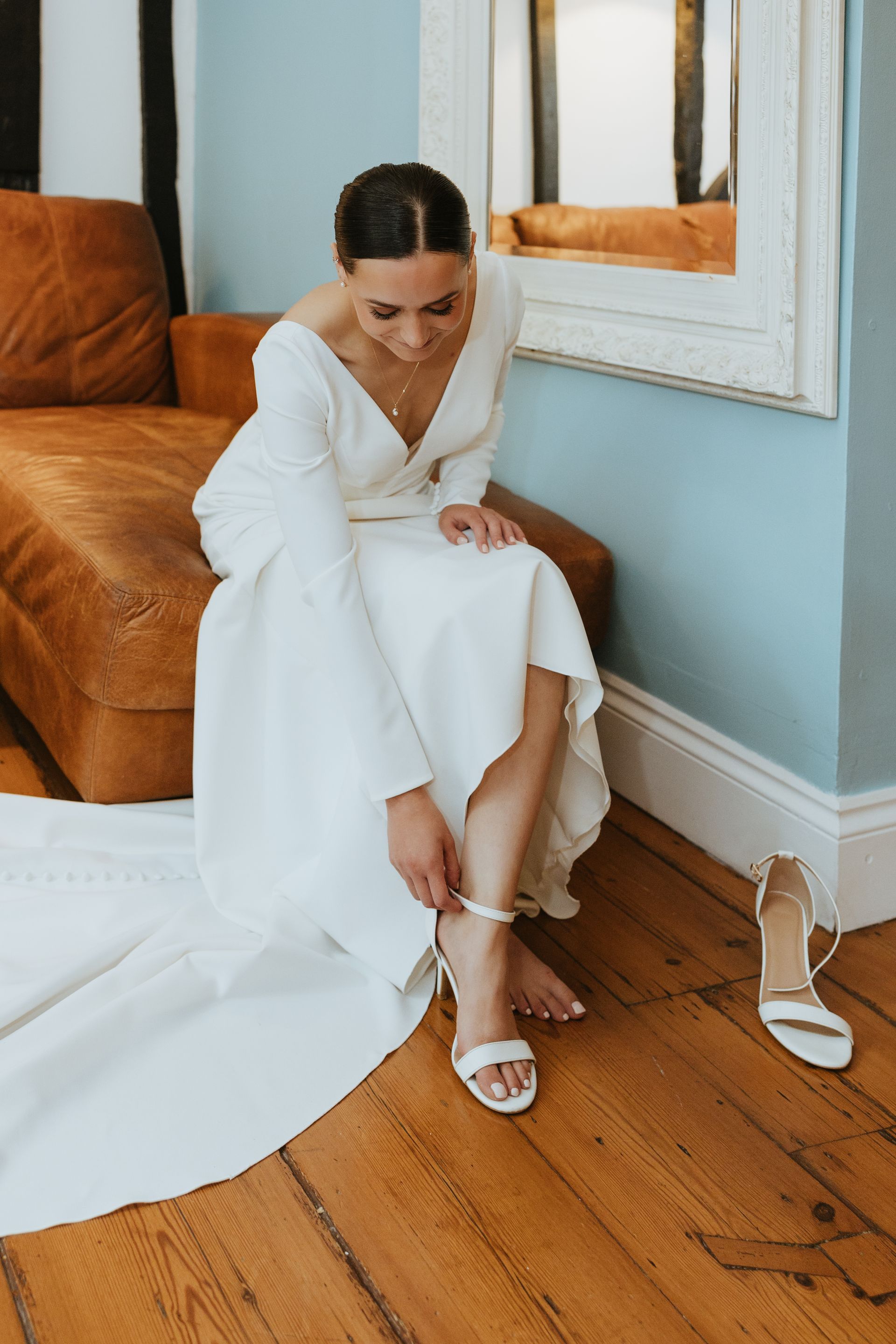 Woman in a white robe fastening a white sandal beside a bed in a sunlit room