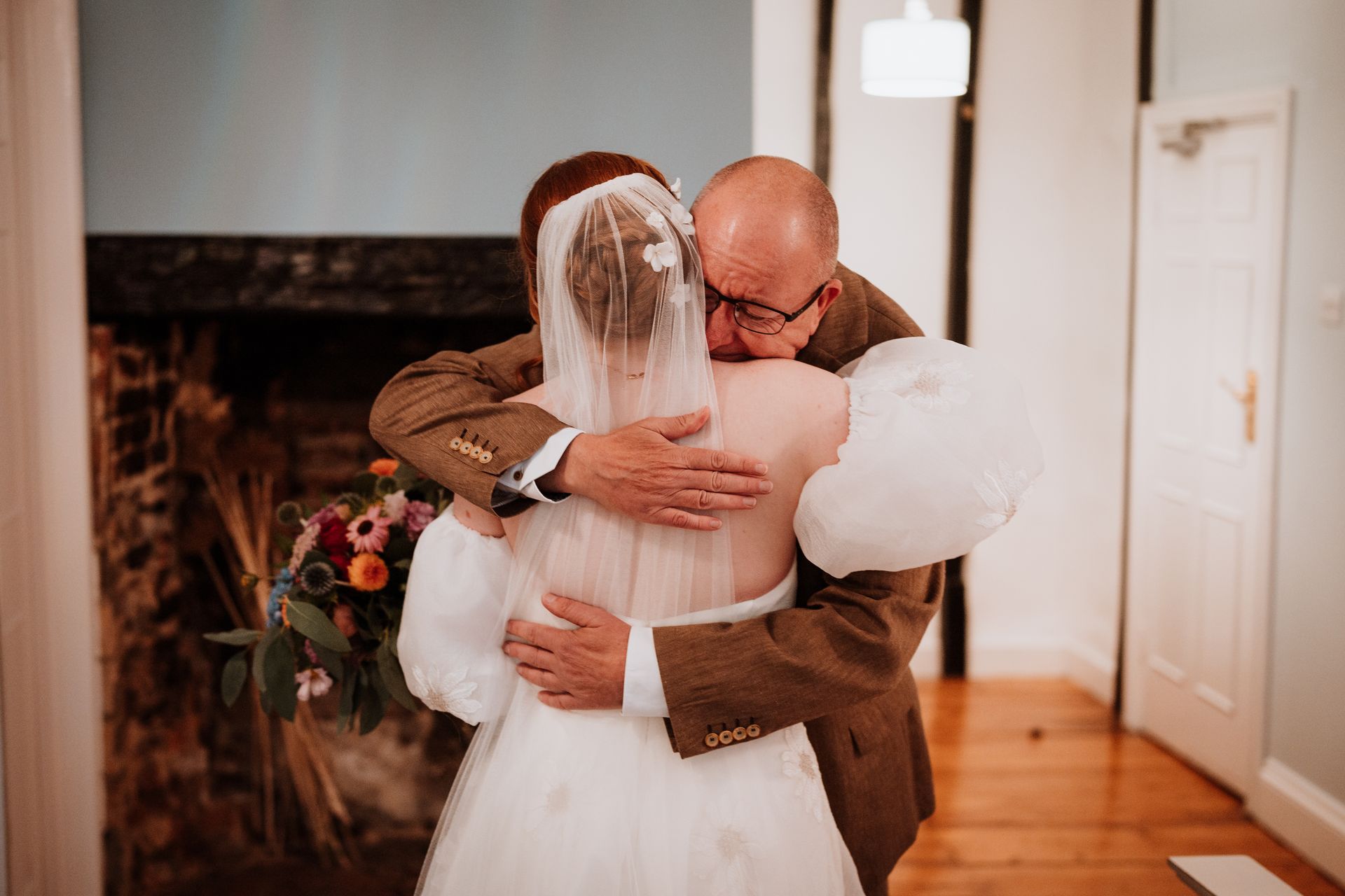 A bride and an older man embrace warmly in a hallway, with the bride in a white dress and veil.