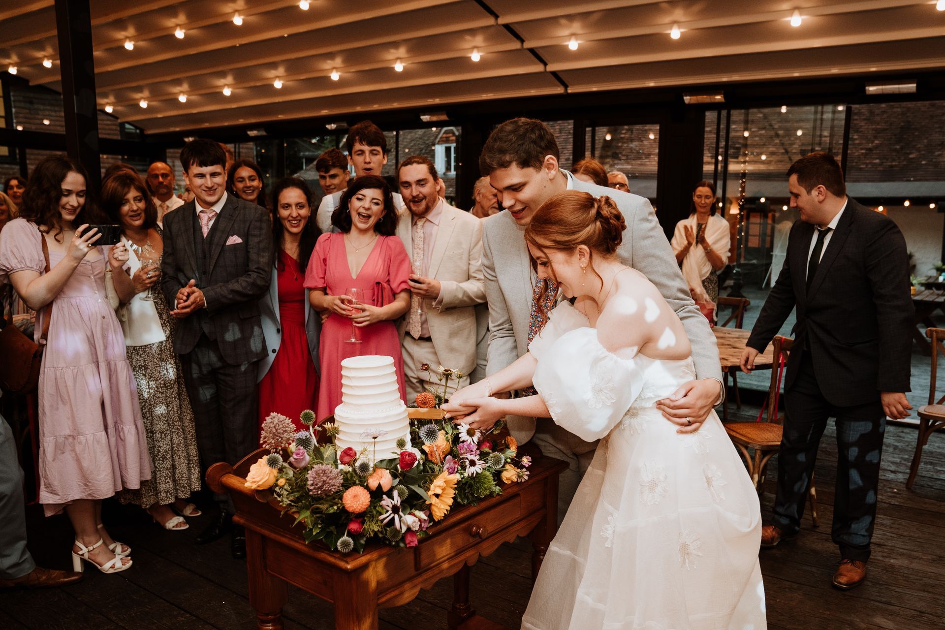 Wedding couple cuts a tiered cake as guests gather around under warm string lights.