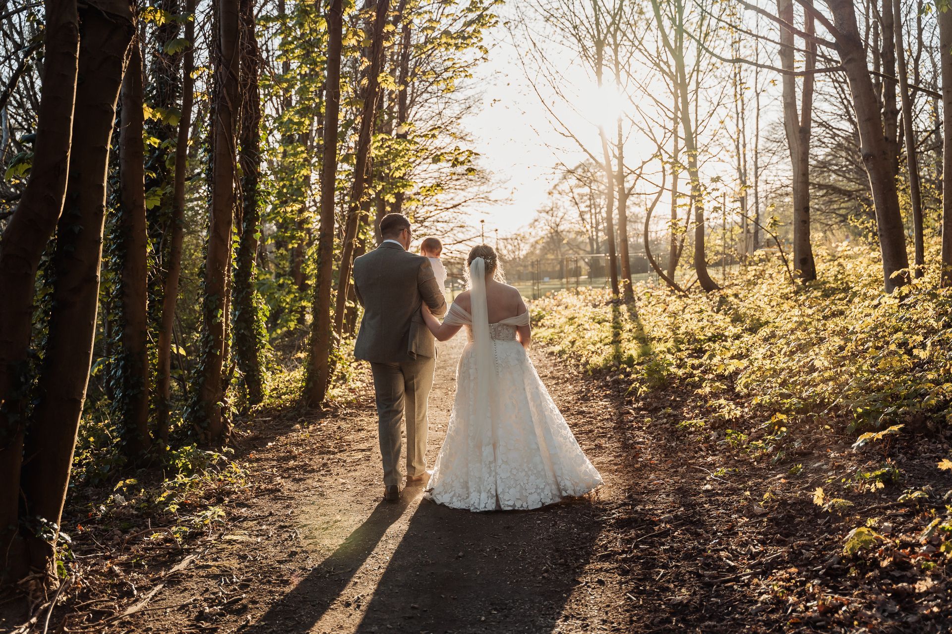 Couple in wedding attire walking hand in hand through a sunlit forest path