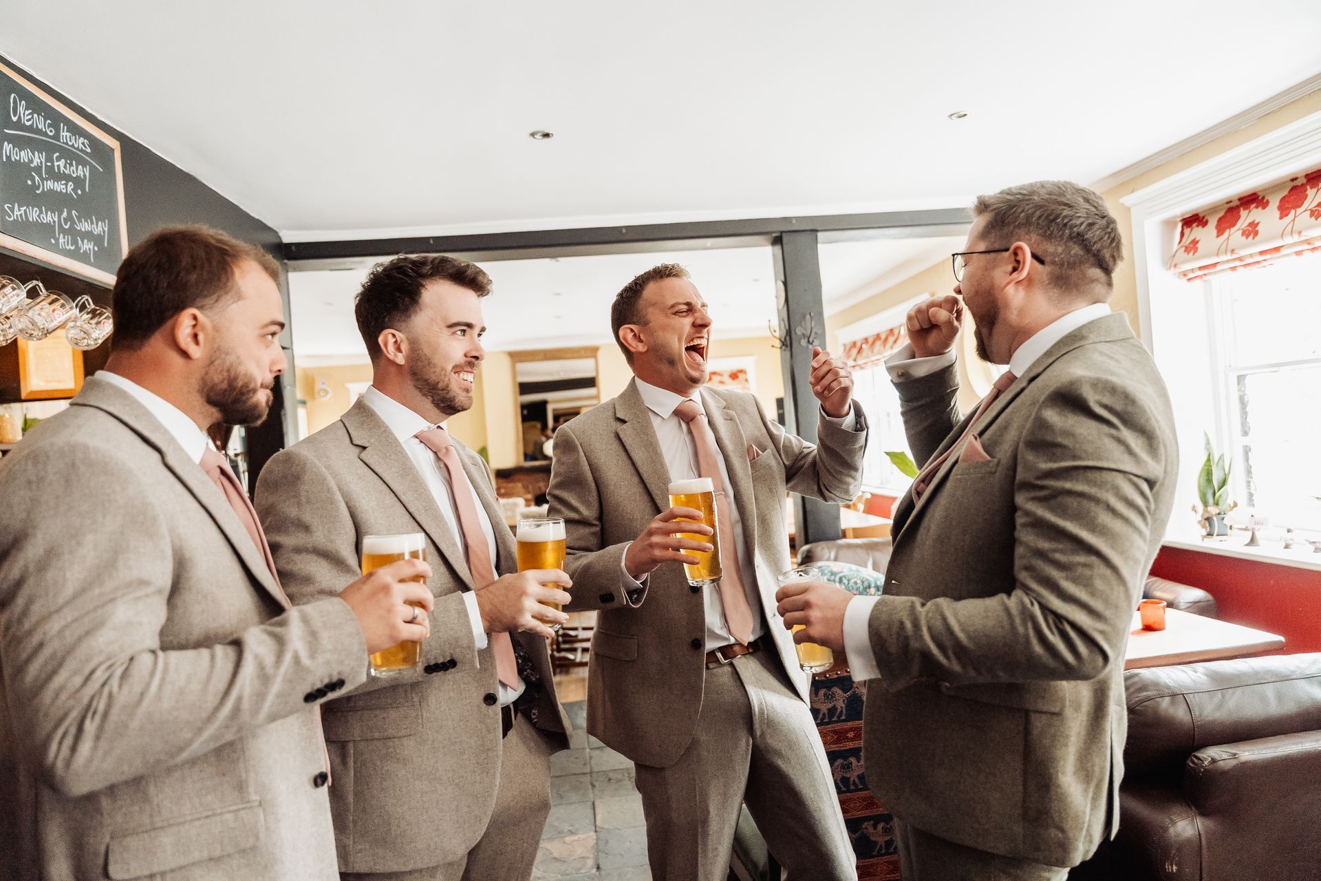 Four men in suits chat and toast with beers at a bright indoor gathering.