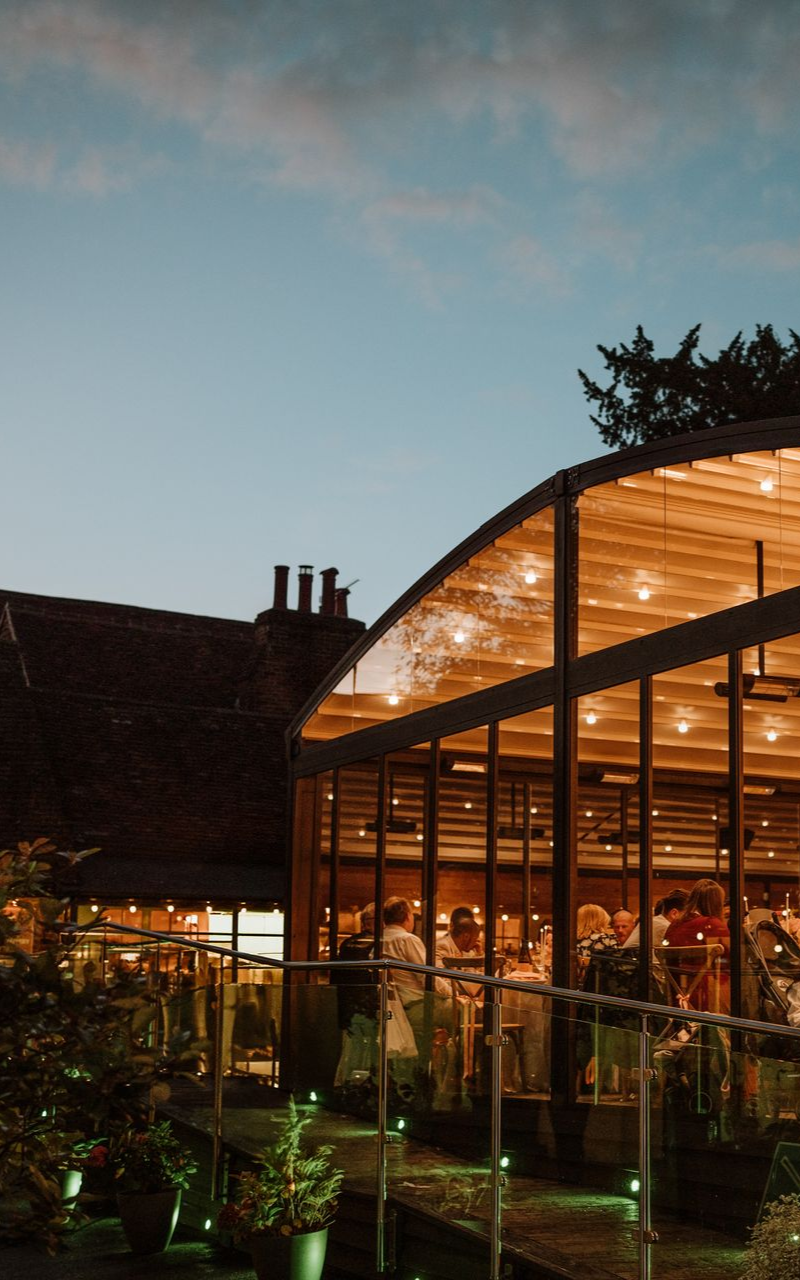 Glass-walled restaurant pavilion glowing warm orange at dusk, with plants and diners inside