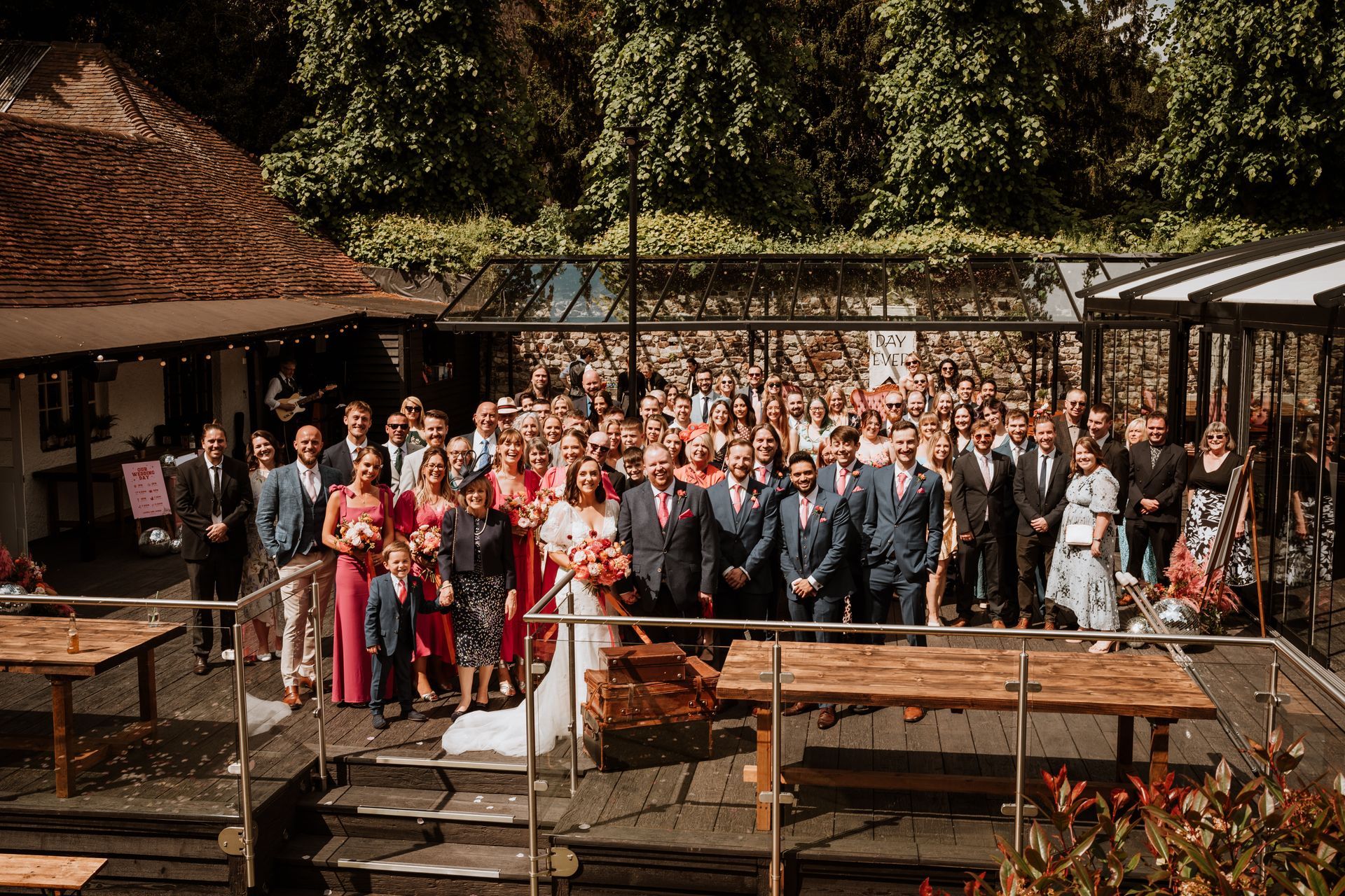 Large wedding party gathered outdoors in front of rustic buildings, with the bride in white and guests in formal attire