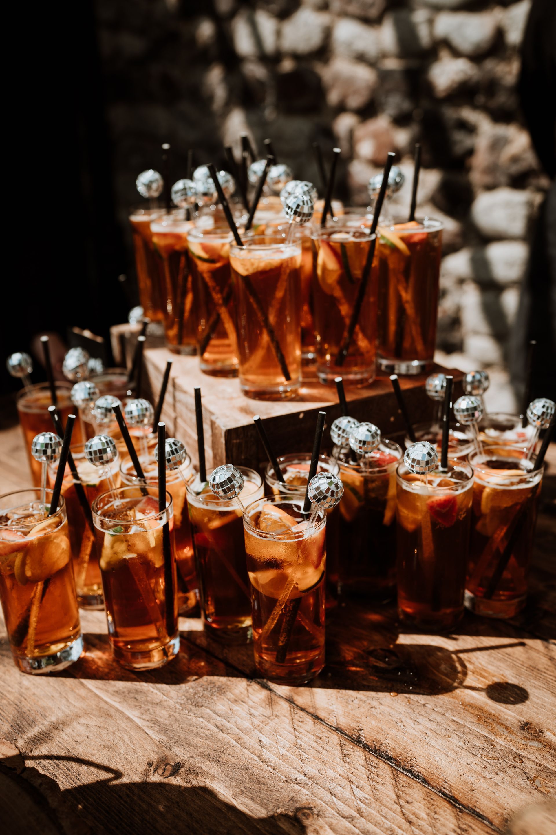 Rows of amber cocktails in small glasses with black straws on a rustic wooden table, outdoors.