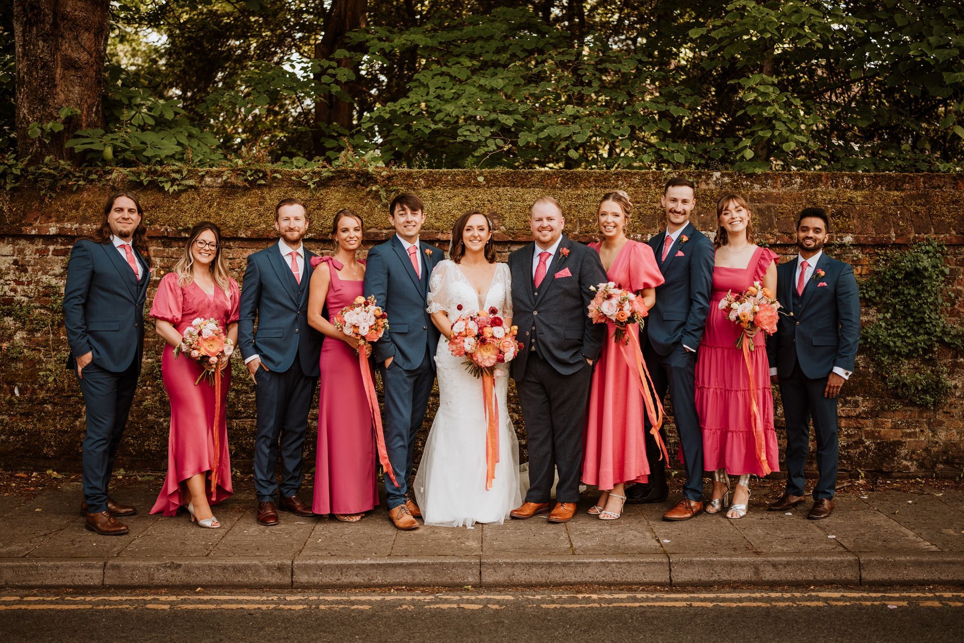 Wedding party posing outdoors in pink and navy attire before a hedge and stone wall.