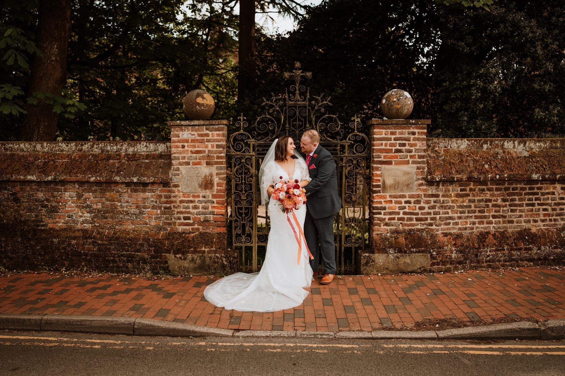 Bride and groom posing by a brick gate in a garden, with the bride holding a bouquet and a white dress