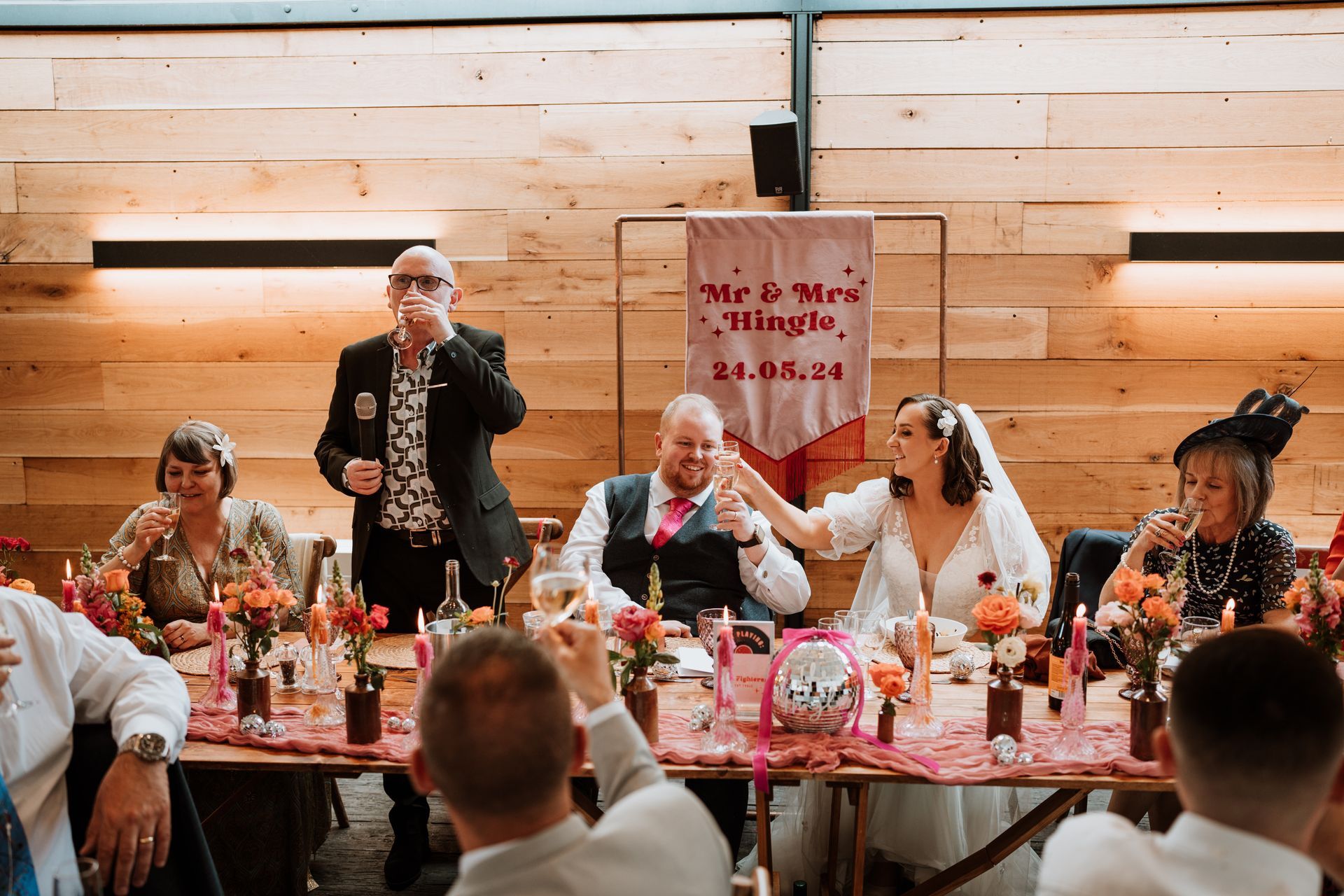 Wedding party at a decorated table; a man stands and raises a glass as guests sit beside a sign and floral centerpieces.