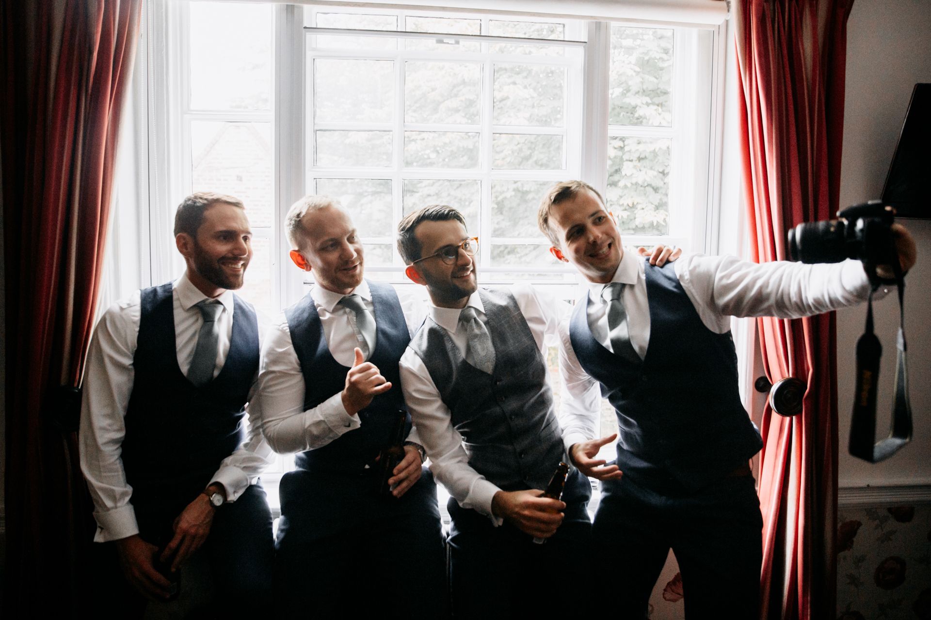 Four men in formal attire take a selfie by a window, smiling and making playful gestures.