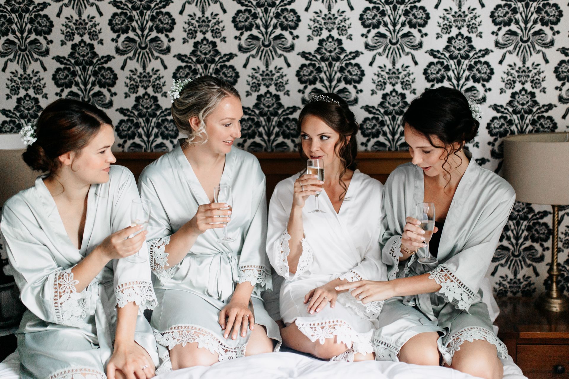 Four women in white robes sit on a bed, chatting and drinking from glasses in a patterned bedroom.