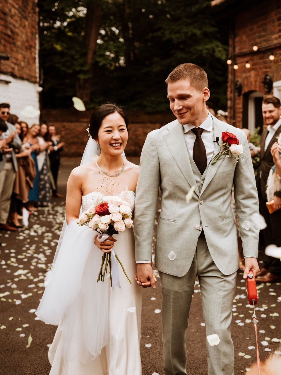Newlyweds walking hand in hand down an outdoor aisle, smiling, with petals falling around them.