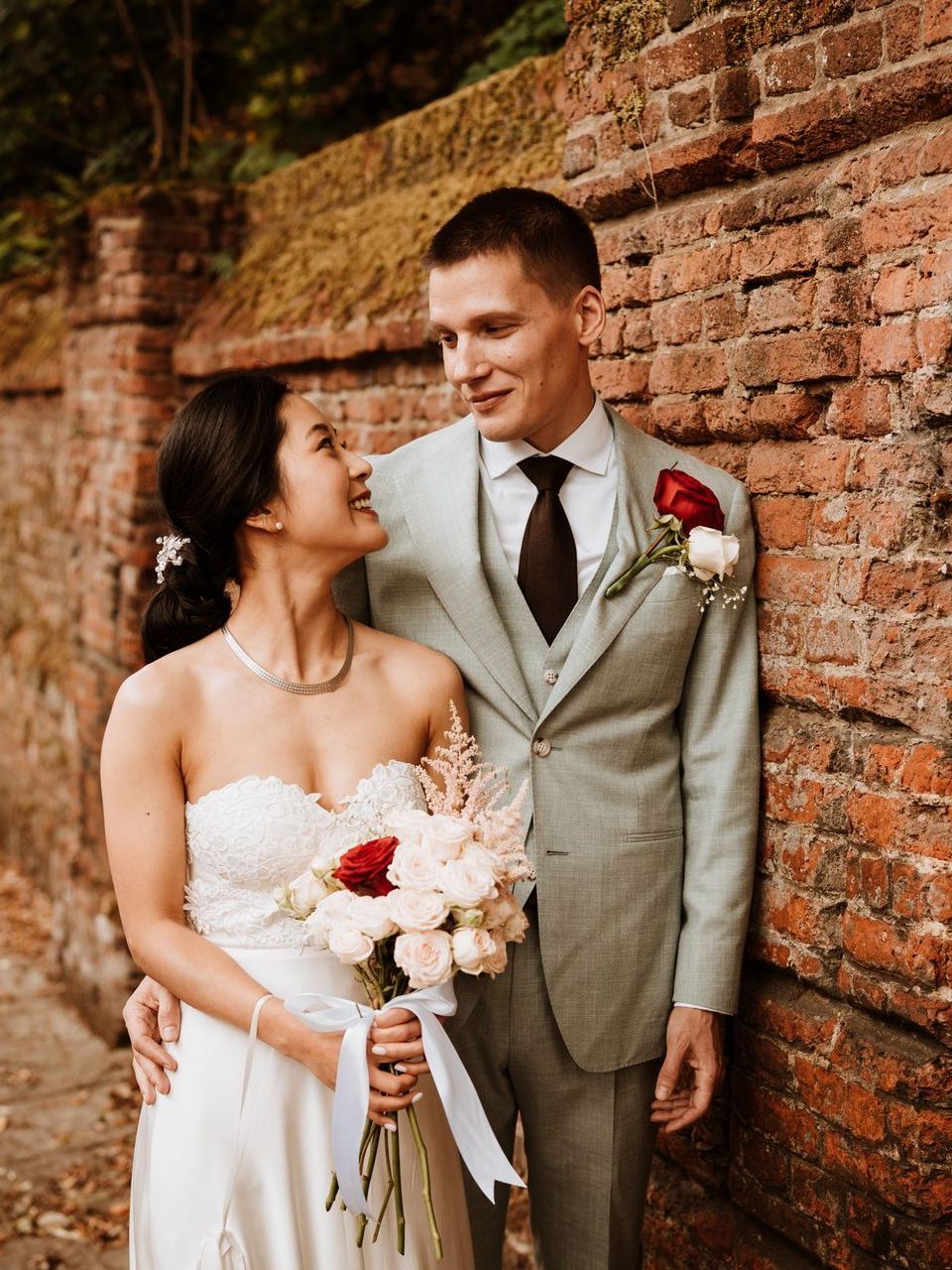 Newlyweds smiling beside a brick wall, bride in white dress holding bouquet, groom in light suit with red boutonniere