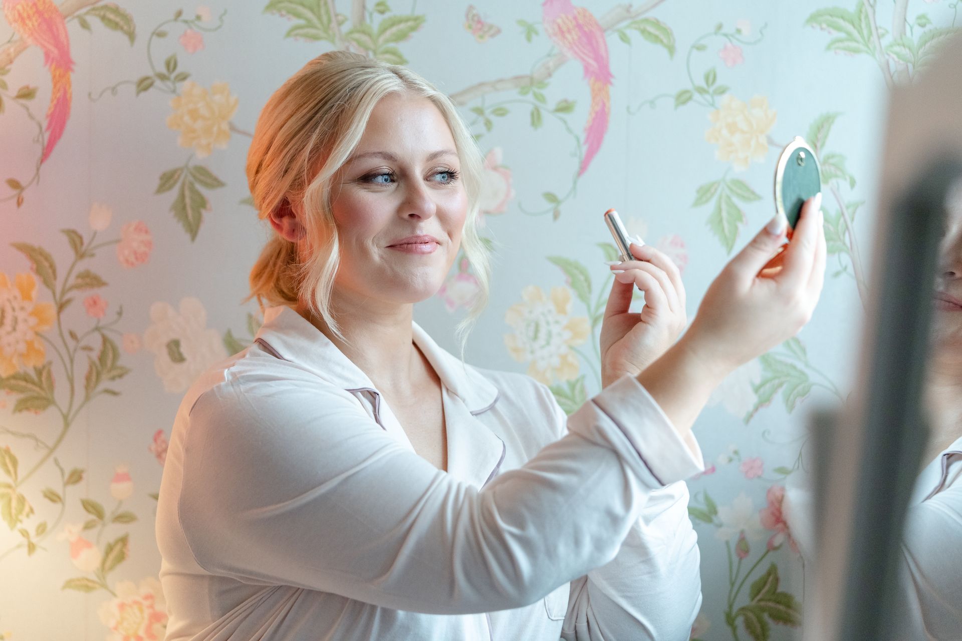 Woman applying makeup in a floral room, holding a compact mirror and brush.