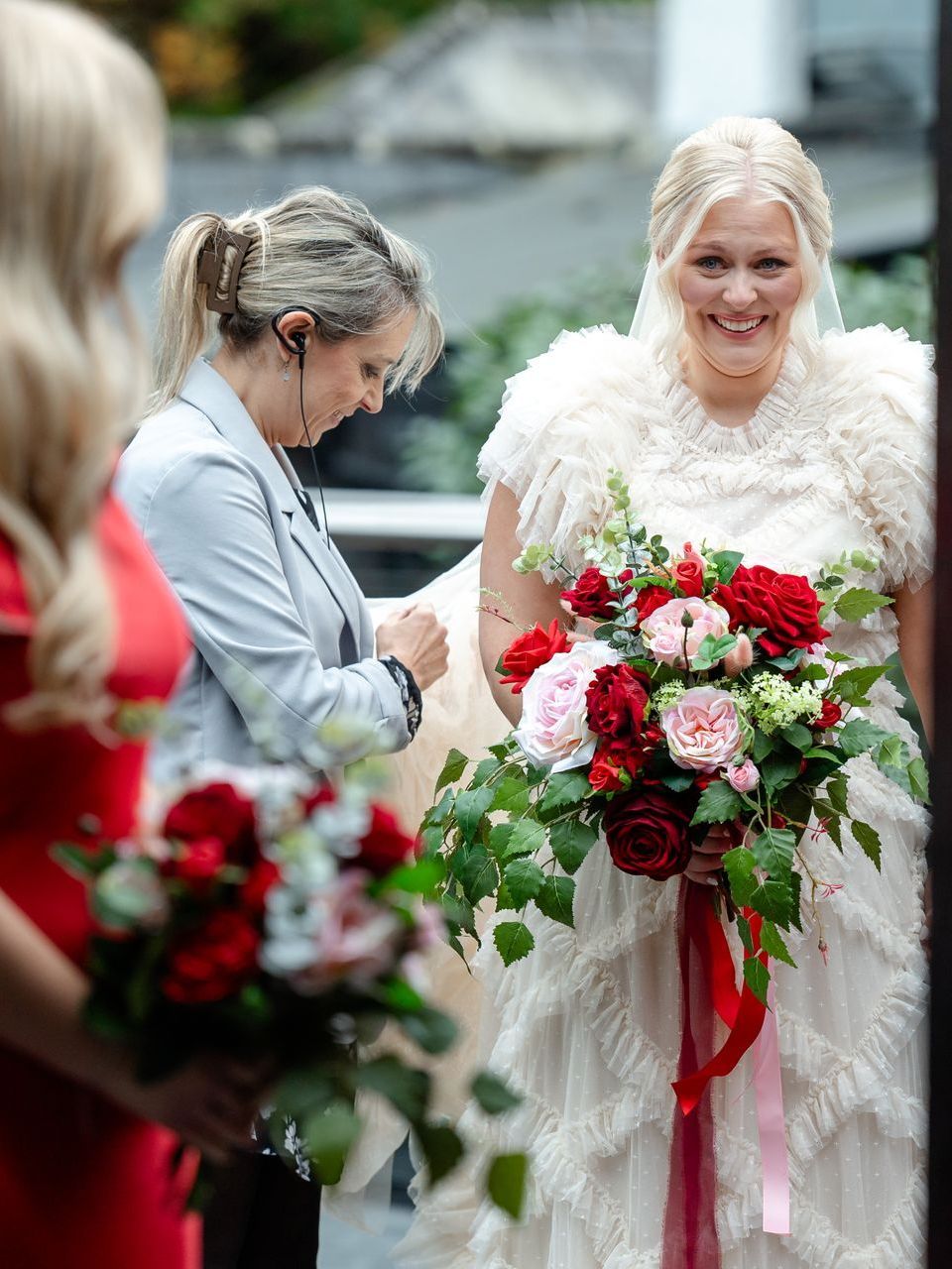 Bride holding a red-and-white bouquet while another woman adjusts her dress at an outdoor wedding ceremony