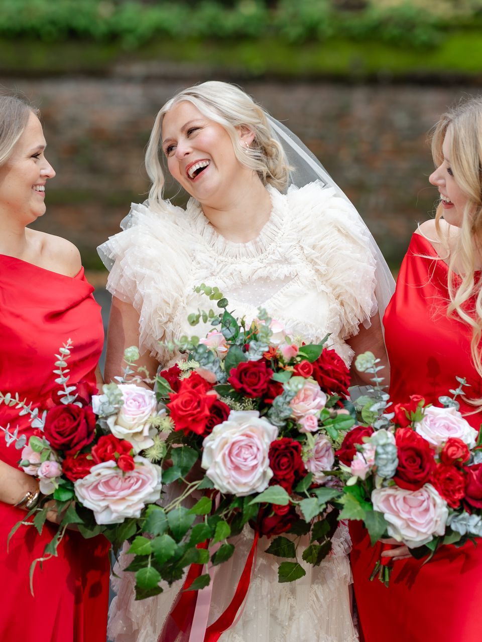 Three women in red dresses holding bouquets, with the center woman in a white dress smiling outdoors