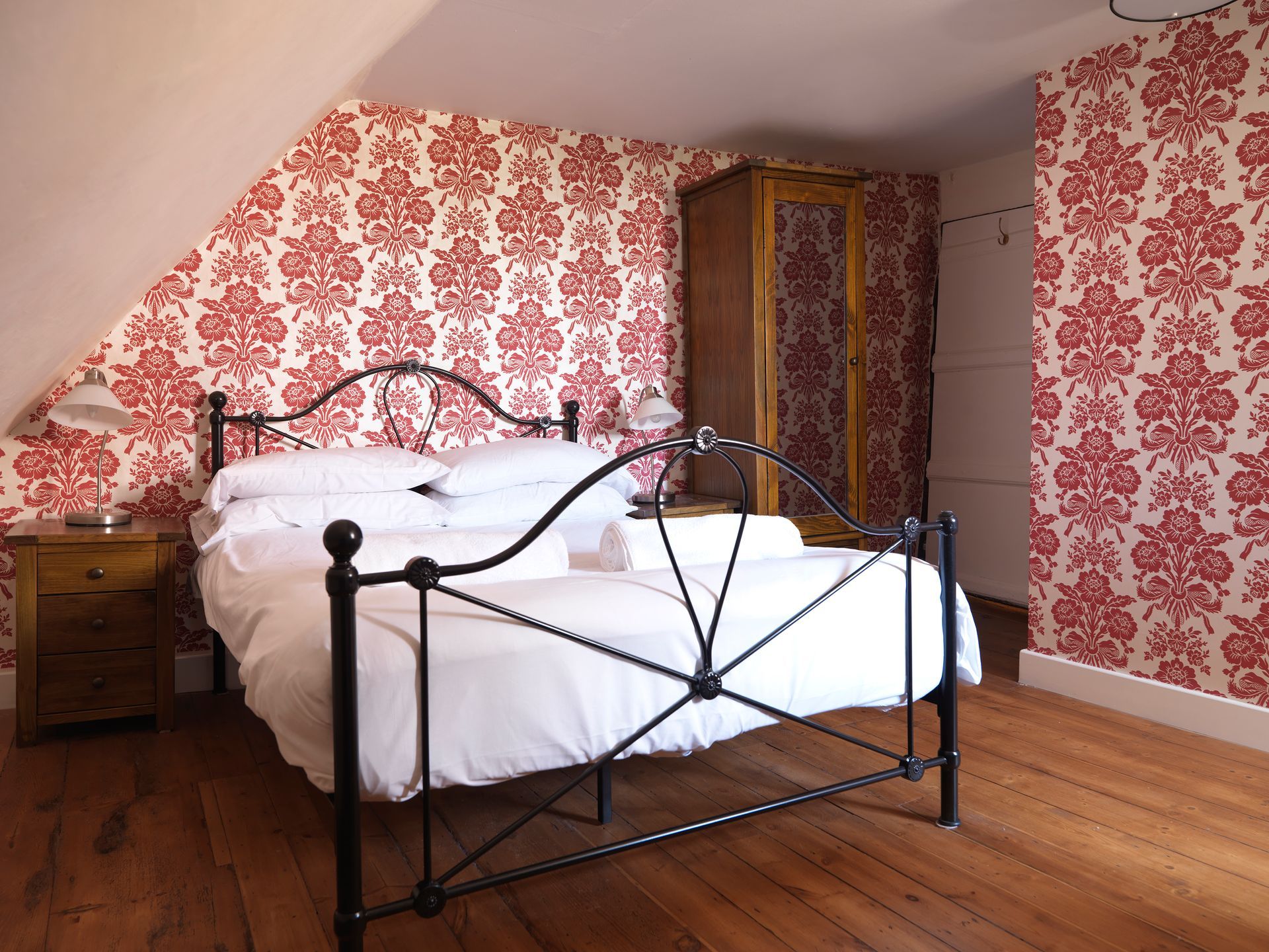 Attic bedroom with red floral wallpaper, wrought-iron bed, wooden floor, and wardrobe.