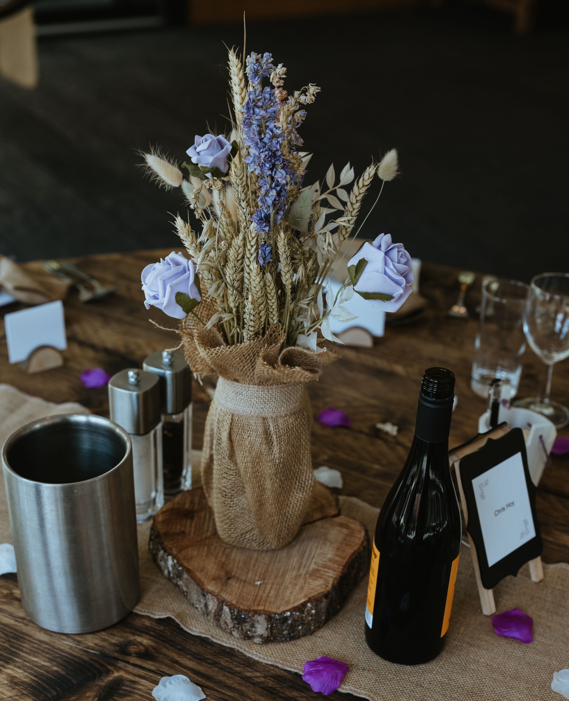 Rustic table centerpiece with dried flowers in a burlap vase, bottles, glasses, and scattered purple petals