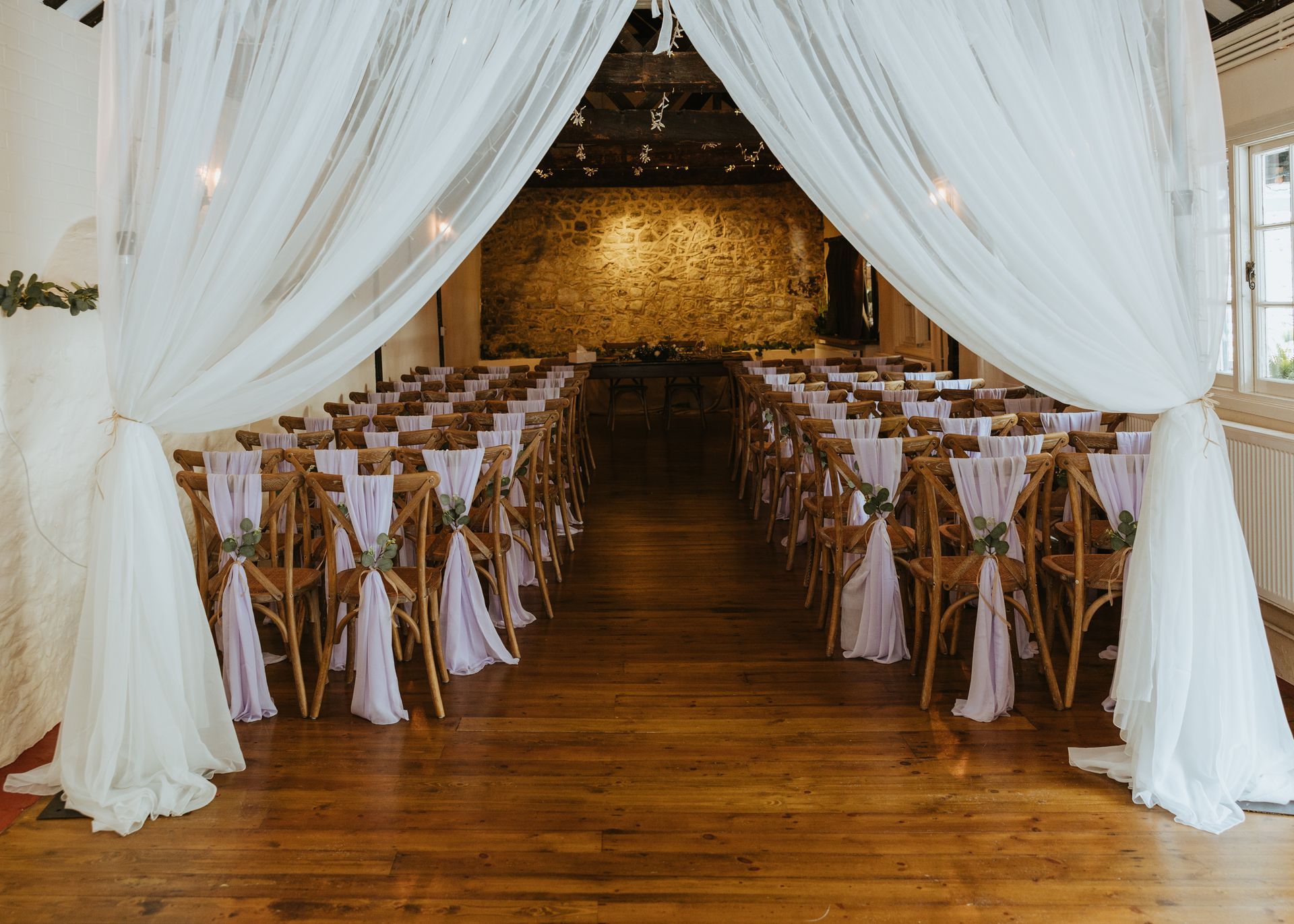 Elegant wedding ceremony setup with white drapes, gold chairs, and a wooden aisle in a rustic hall