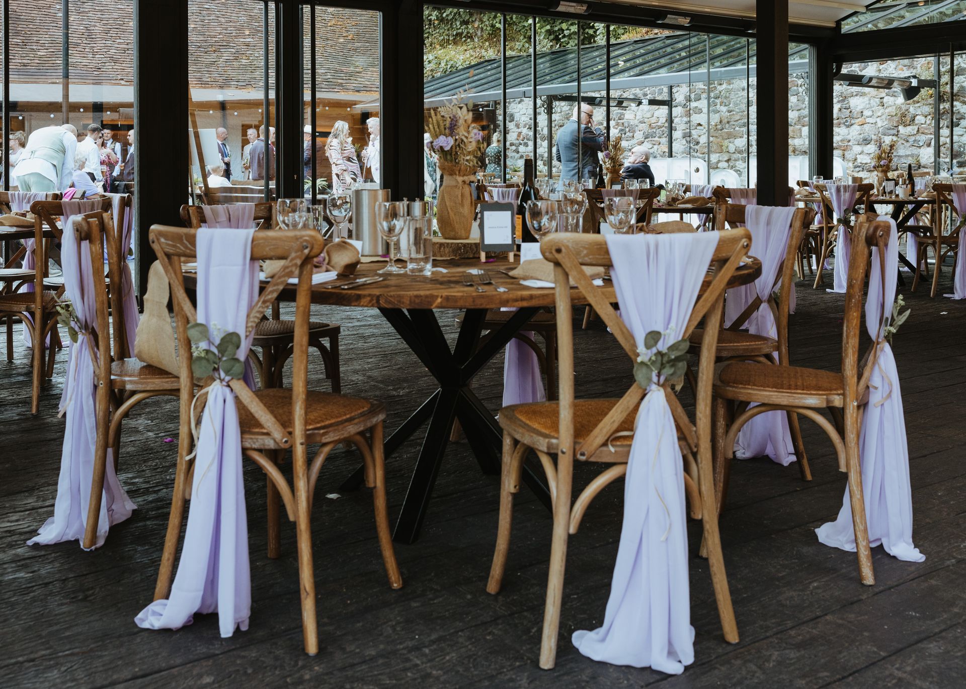 Decorated outdoor dining table with wooden chairs, white drapes, and floral centerpieces under a covered patio