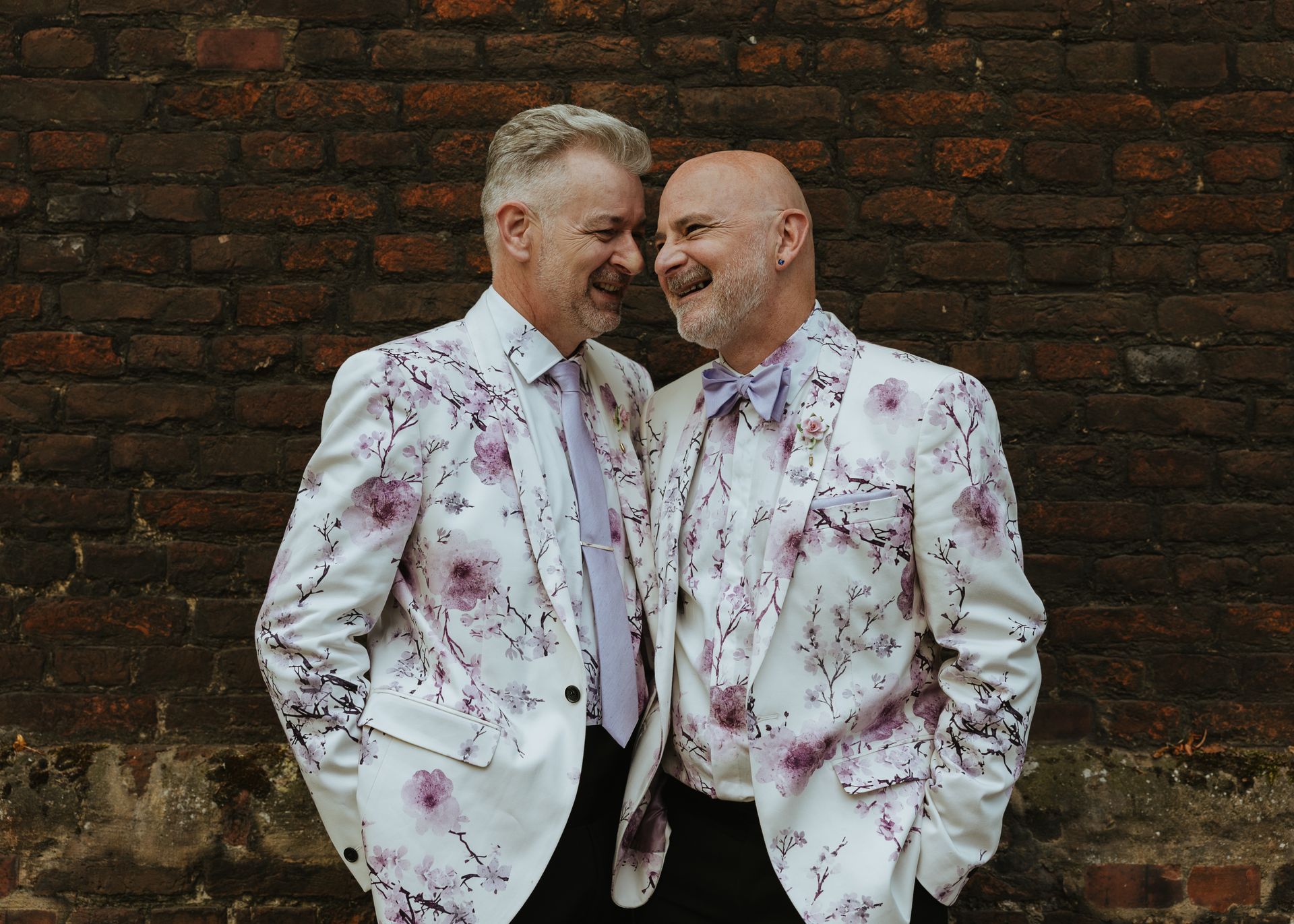 Two men in white floral jackets smiling against a brick wall.