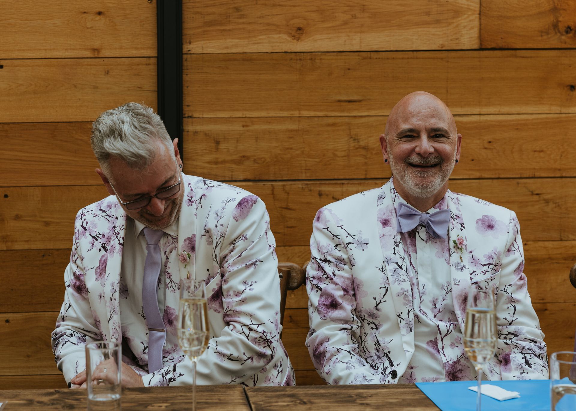 Two men in white floral suits seated at a table against a wooden wall, one smiling and one looking down.