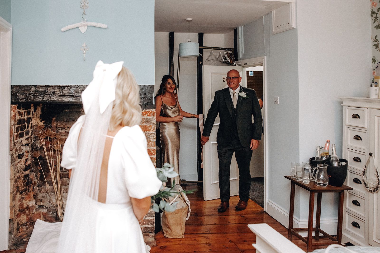 Bride entering a living room as a man in a suit greets her, with another woman behind him