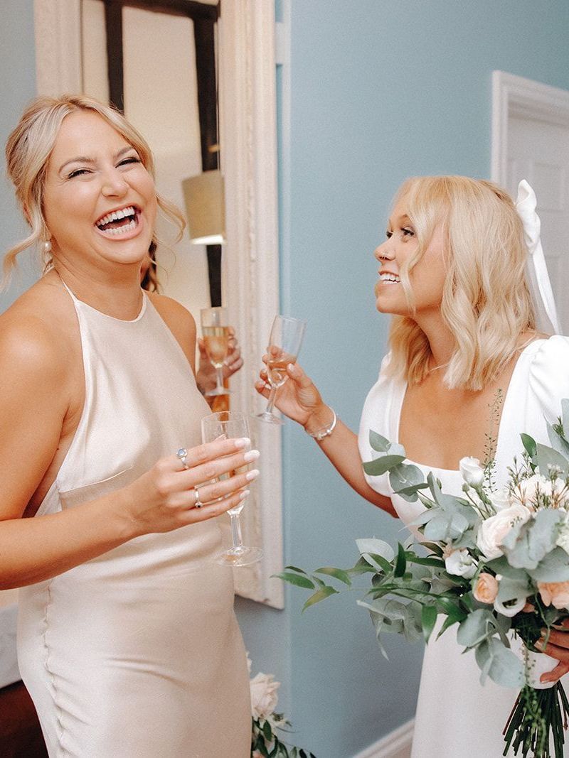 Two women in light dresses smiling and toasting with champagne in a bright room, one holding white flowers