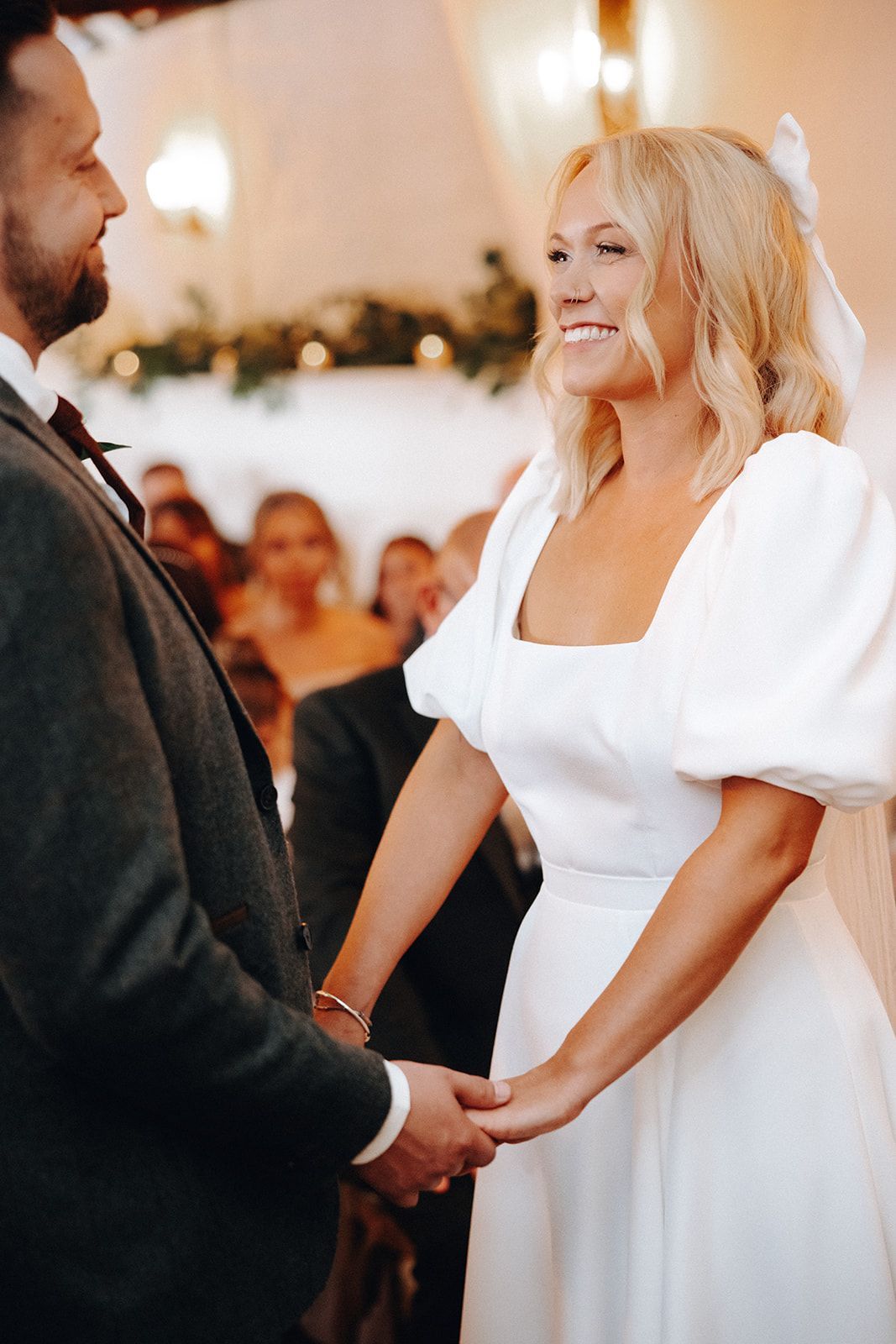 Couple holding hands during wedding vows, bride in white dress smiling indoors with guests blurred behind them