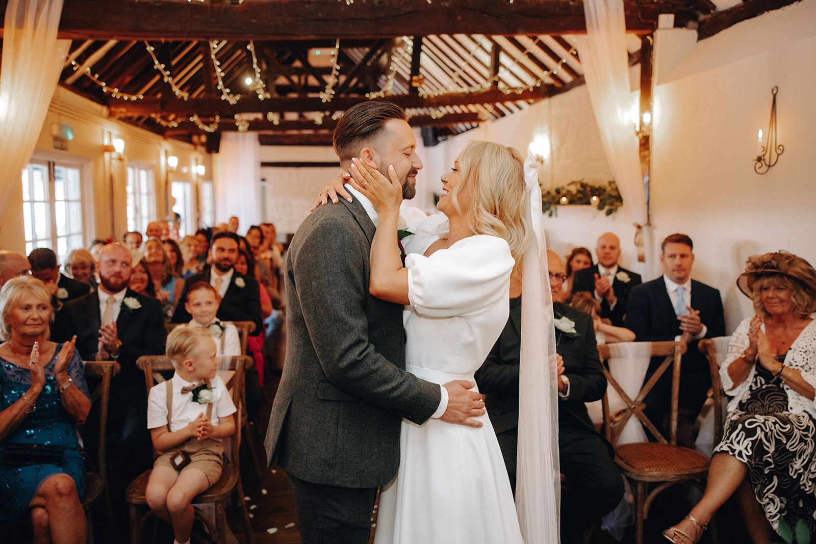 Couple dancing at a rustic indoor wedding ceremony, guests seated around them under warm lights.