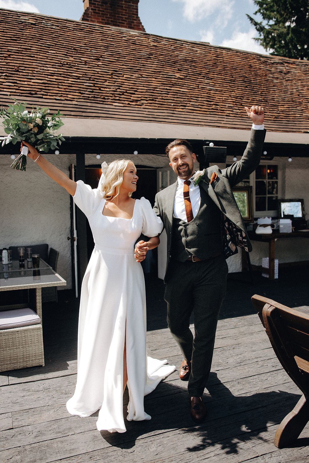 Two people in wedding attire raise their arms and smile outdoors beside a rustic building.