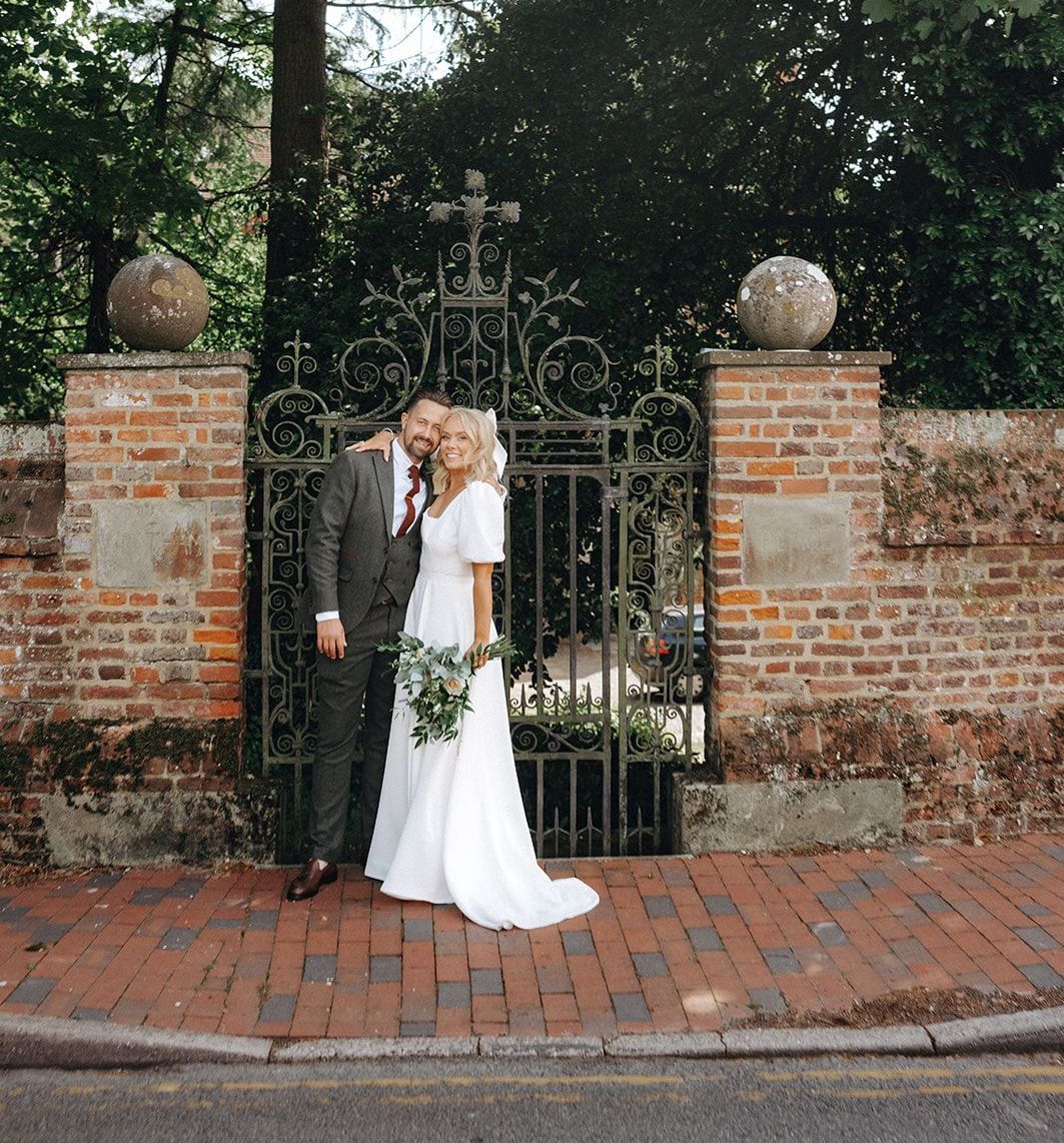 Couple standing by an ornate iron gate, with brick pillars and trees in the background.