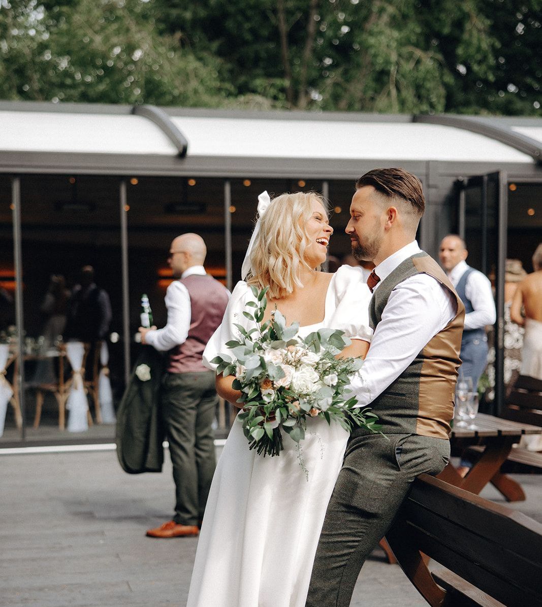 Wedding couple embracing outdoors, bride in white dress holding bouquet, groom in vest at a reception venue