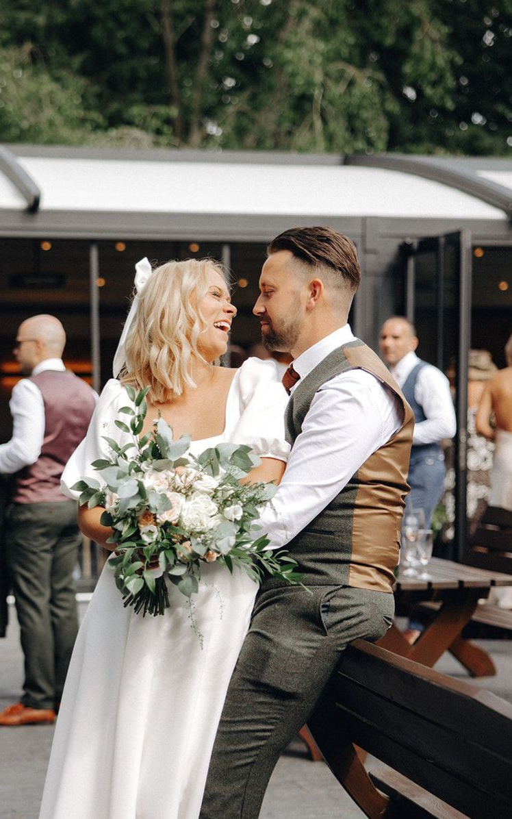 Bride and groom sit together outdoors, smiling and holding a bouquet at a wedding reception