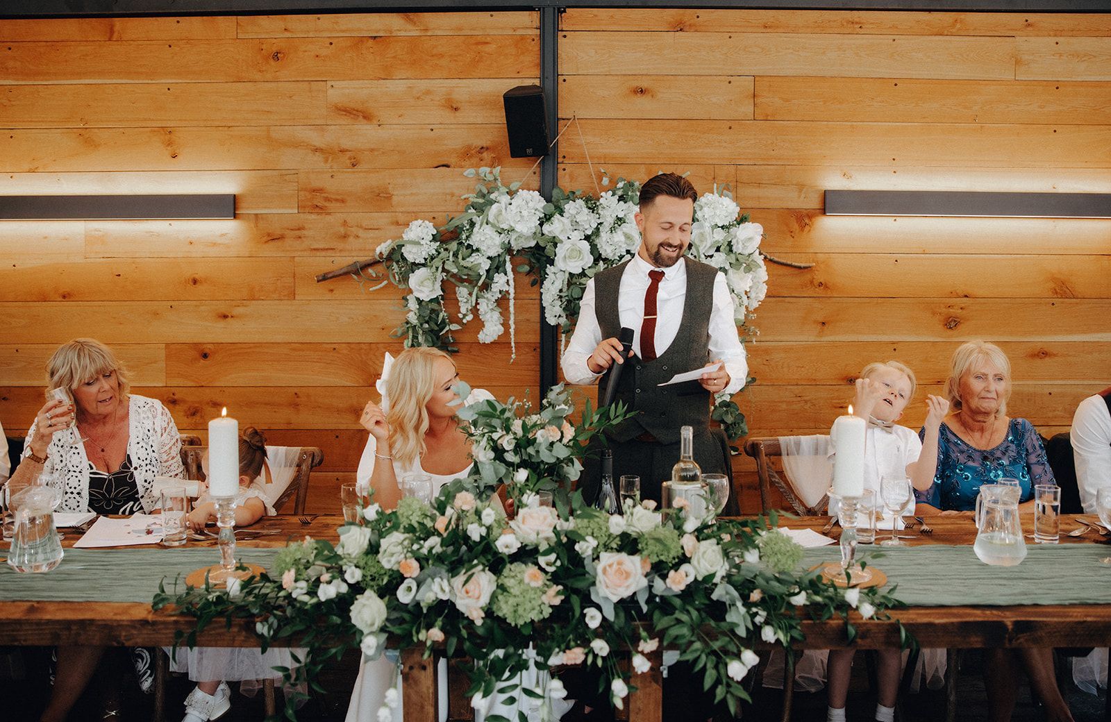 Speaker at floral-decorated wedding reception table with guests seated on either side