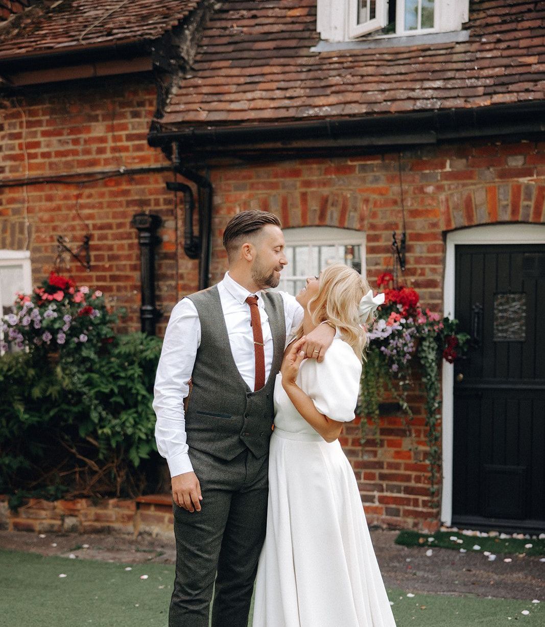 Two people in wedding attire standing outside a brick house, looking at each other lovingly.