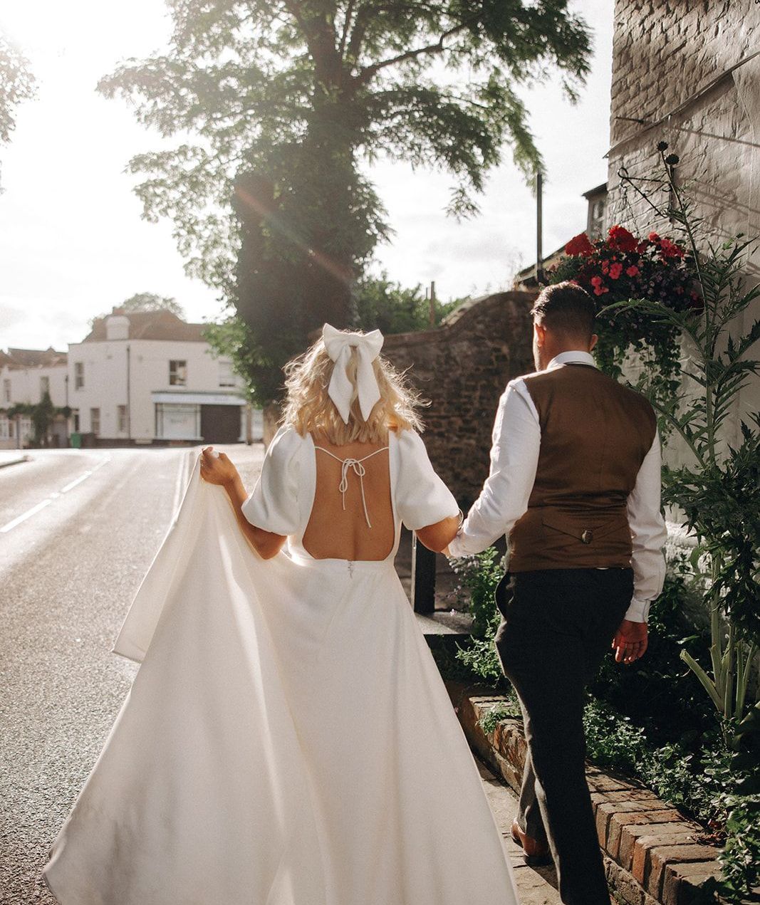 Bride and groom walking hand in hand down a sunlit street in wedding attire