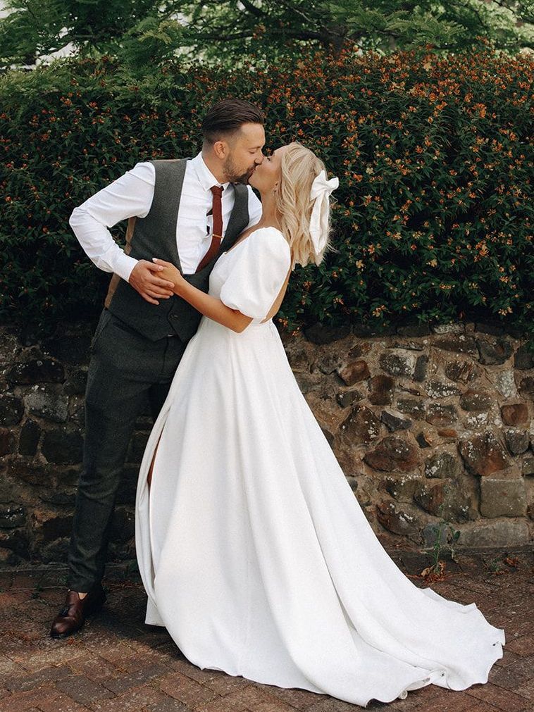 Bride and groom kissing beside a stone wall with greenery behind them
