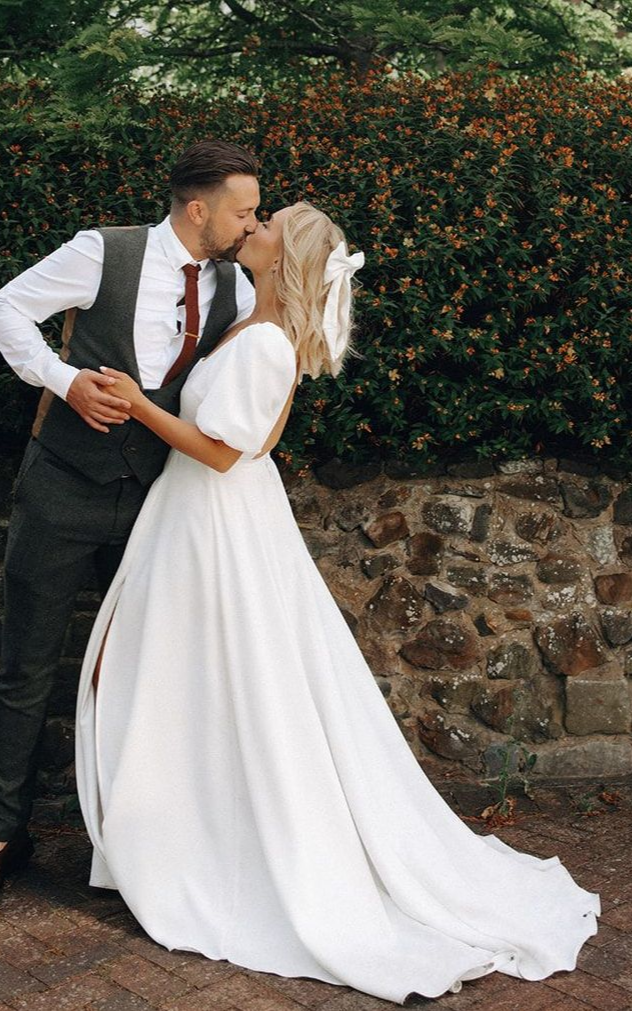 Bride and groom kissing beside a stone wall, bride in a flowing white gown and groom in a black vest