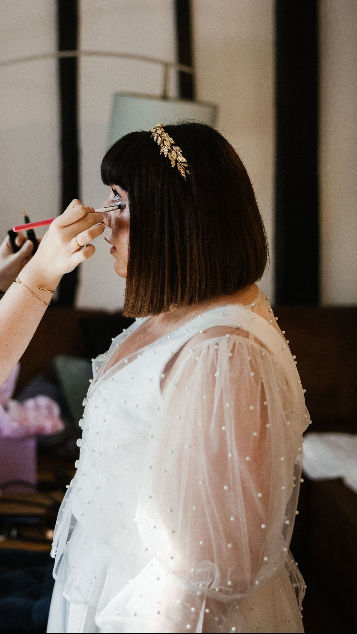 Person in a white dress getting makeup applied indoors, with a floral hair accessory.