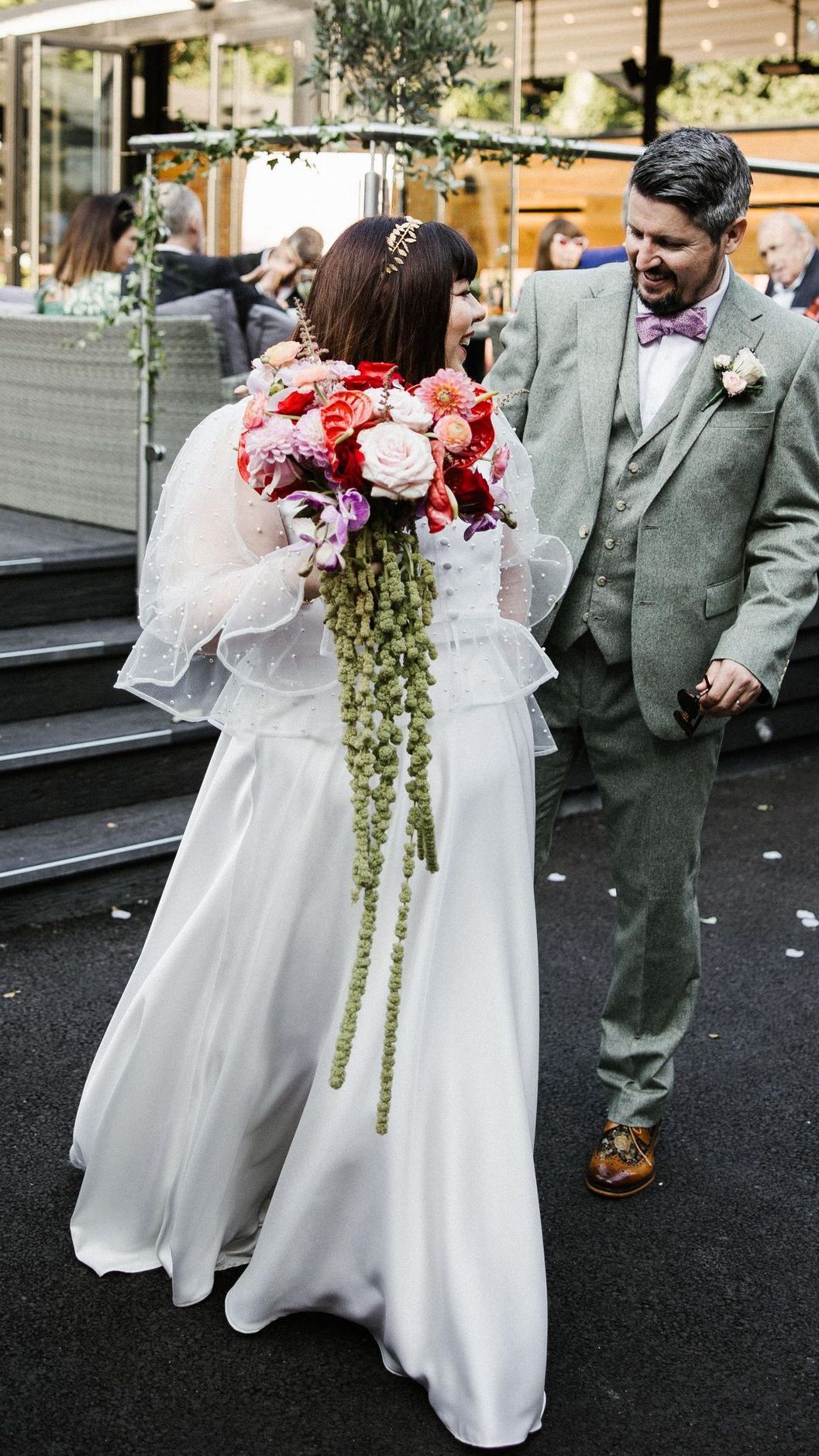 Bride in white gown holding a large pink bouquet beside groom in gray suit outdoors