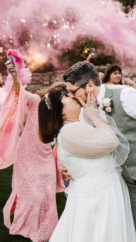 Wedding couple kissing outdoors amid pink smoke and guests celebrating nearby