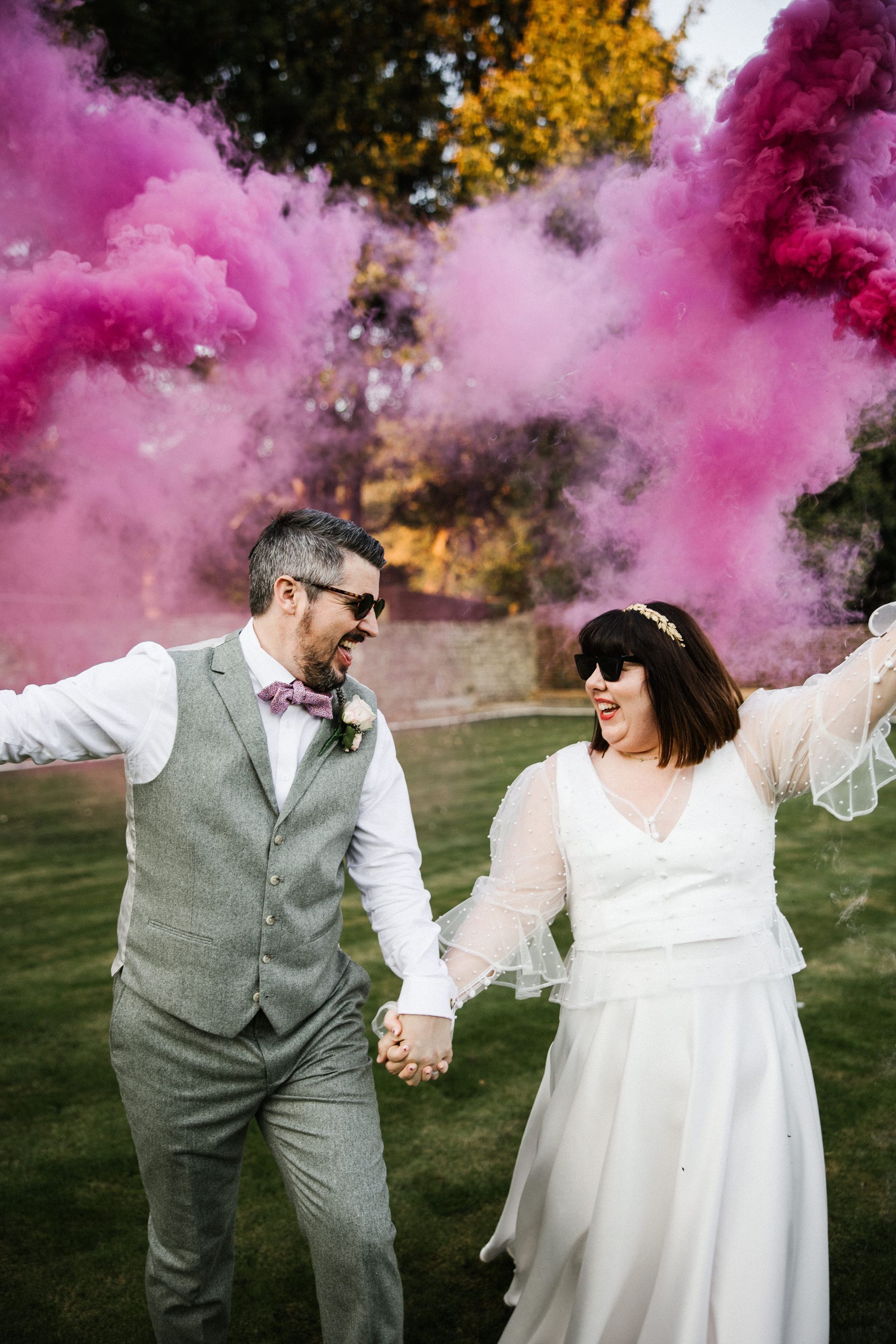 Couple in wedding attire holding hands amid pink smoke at an outdoor celebration