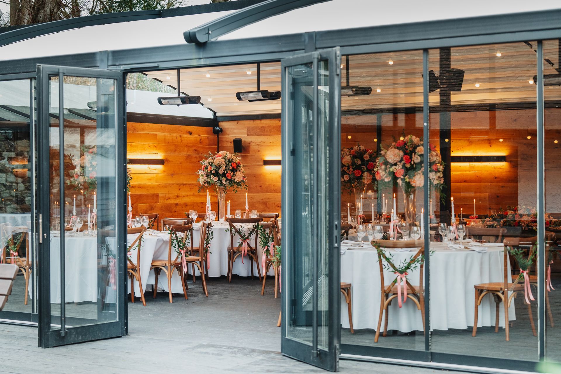 Glass-walled banquet hall with round tables, white linens, floral centerpieces, and warm lighting