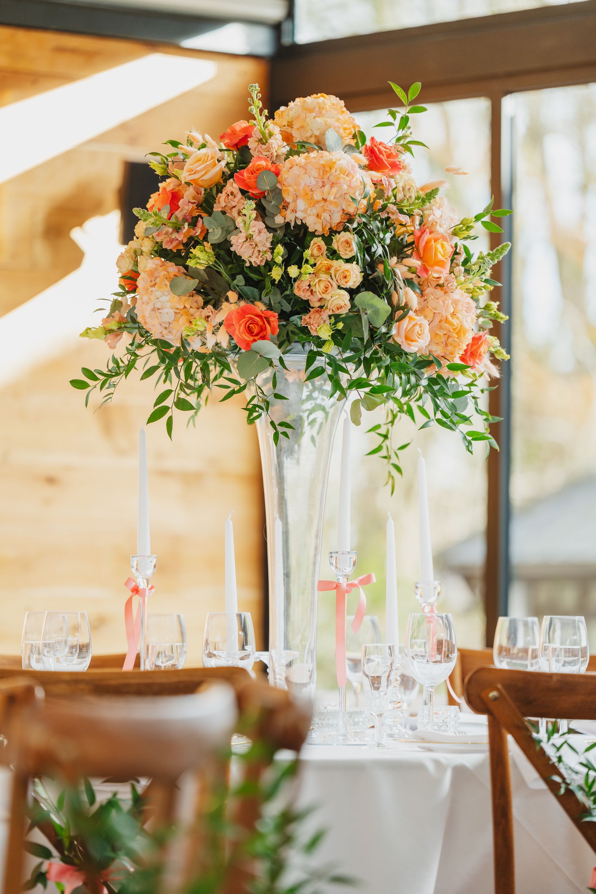 Elegant table setting with tall floral arrangement, peach and coral flowers, candles, and clear glassware.