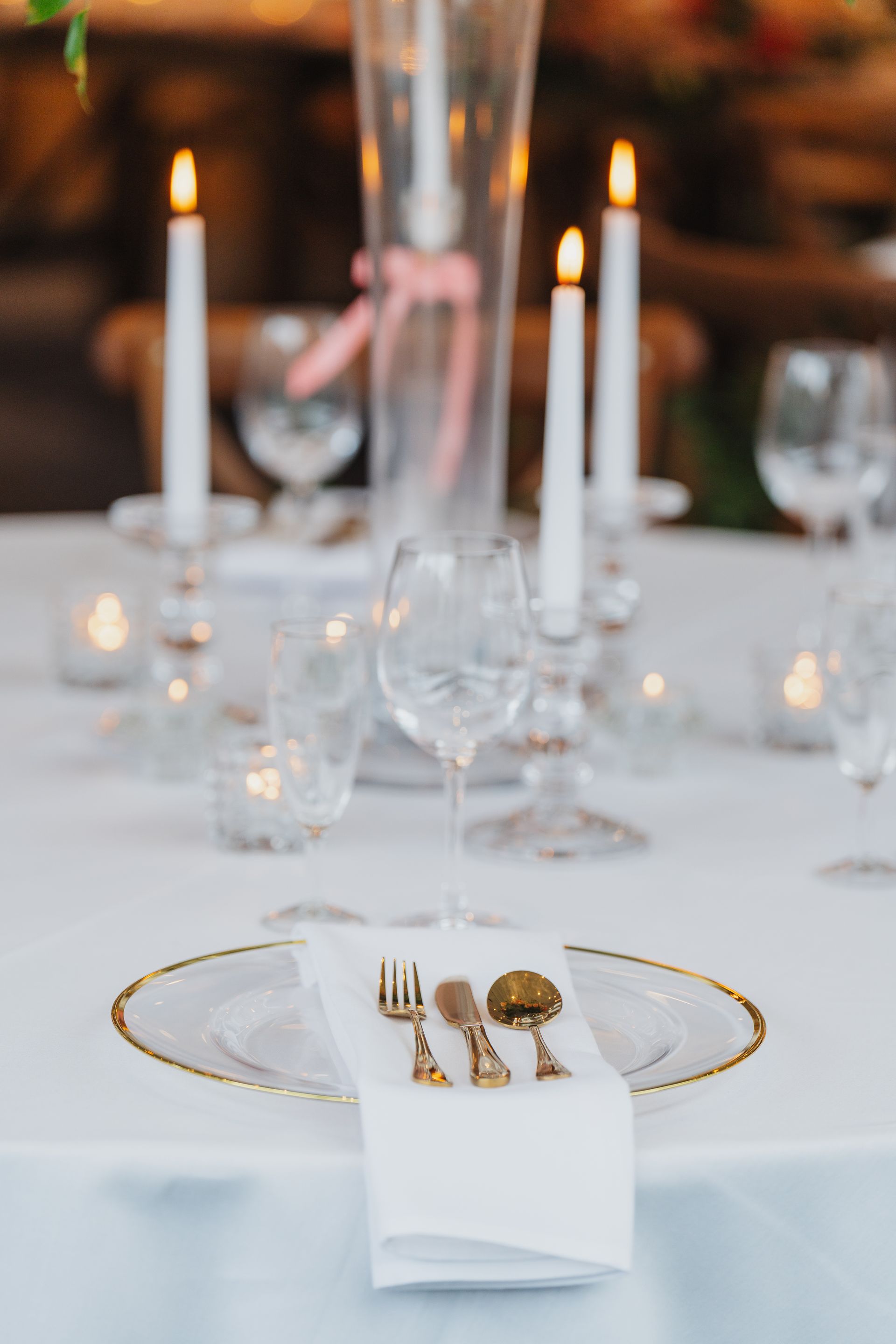 Elegant wedding table setting with gold-rimmed plates, white napkin, ornate silverware, and lit candles.