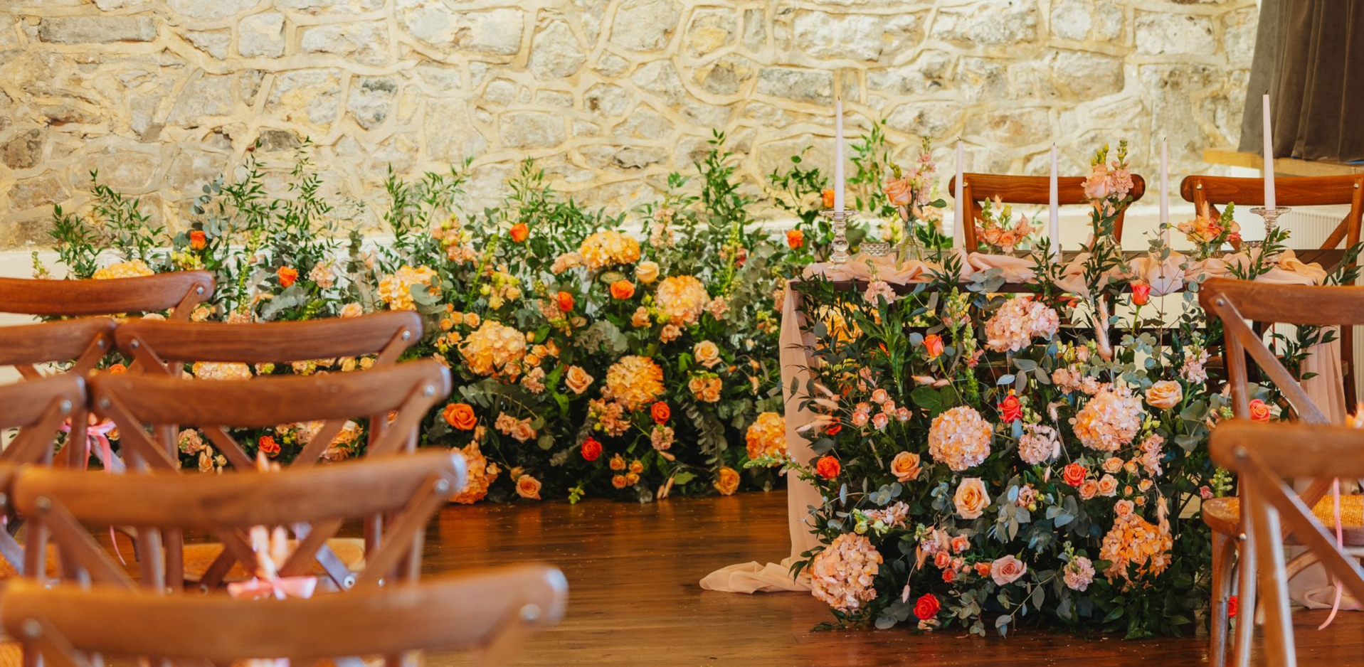 Wedding ceremony setup with wooden chairs facing a floral arrangement and stone wall backdrop.