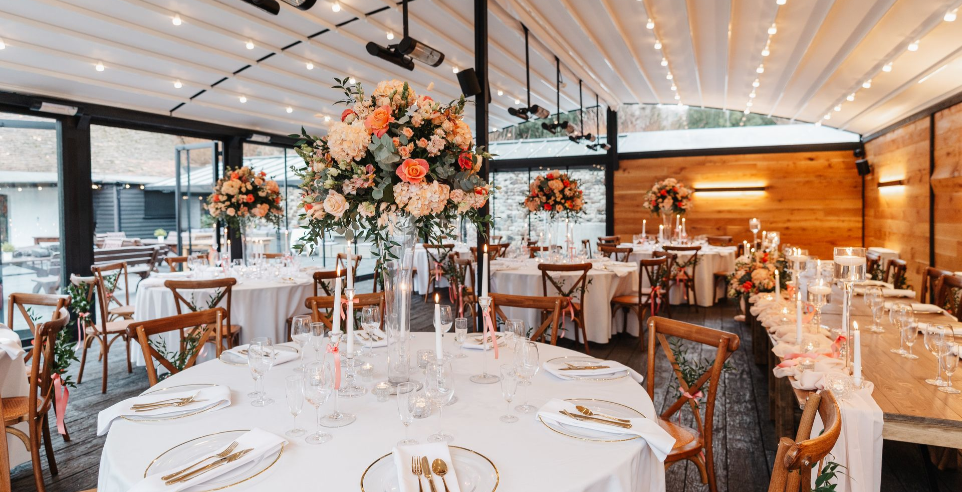 Wedding reception: round tables with floral centerpieces, candles, wooden chairs, and a retractable roof.