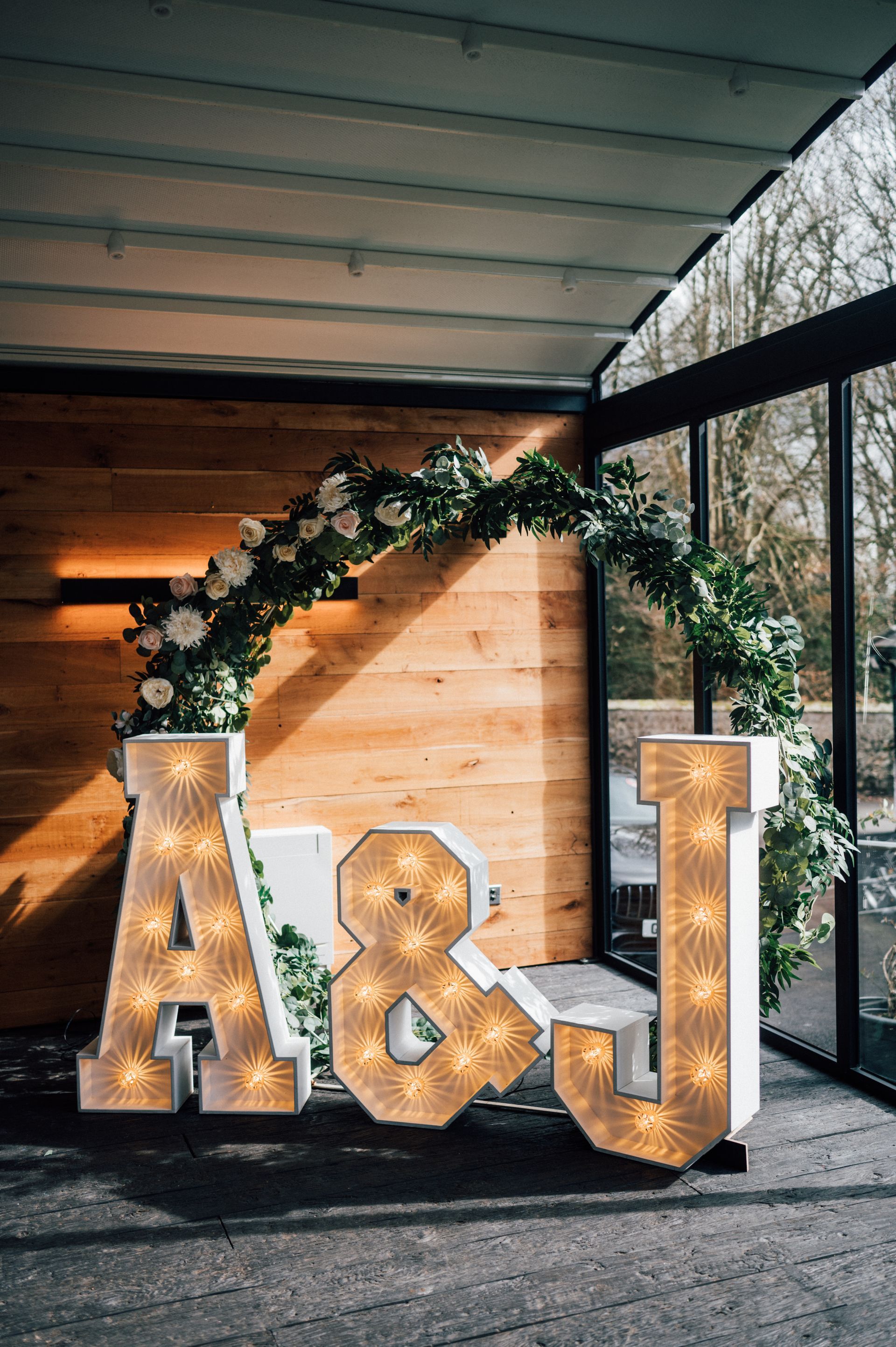 Large glittering “A & J” letters with a greenery arch at an outdoor event backdrop